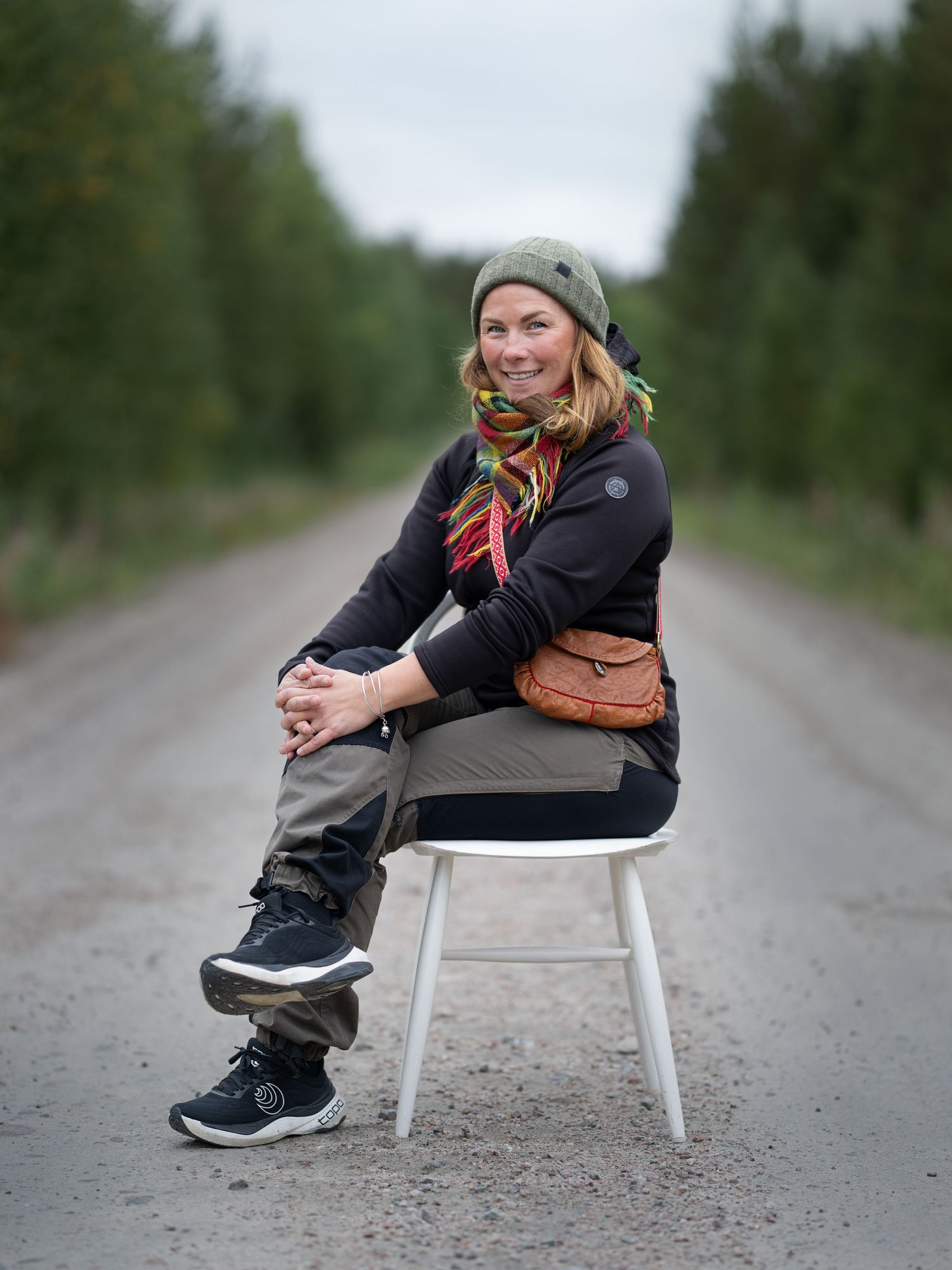 Person sitting casually on a white chair on a rural dirt road wearing a beanie and black jacket.