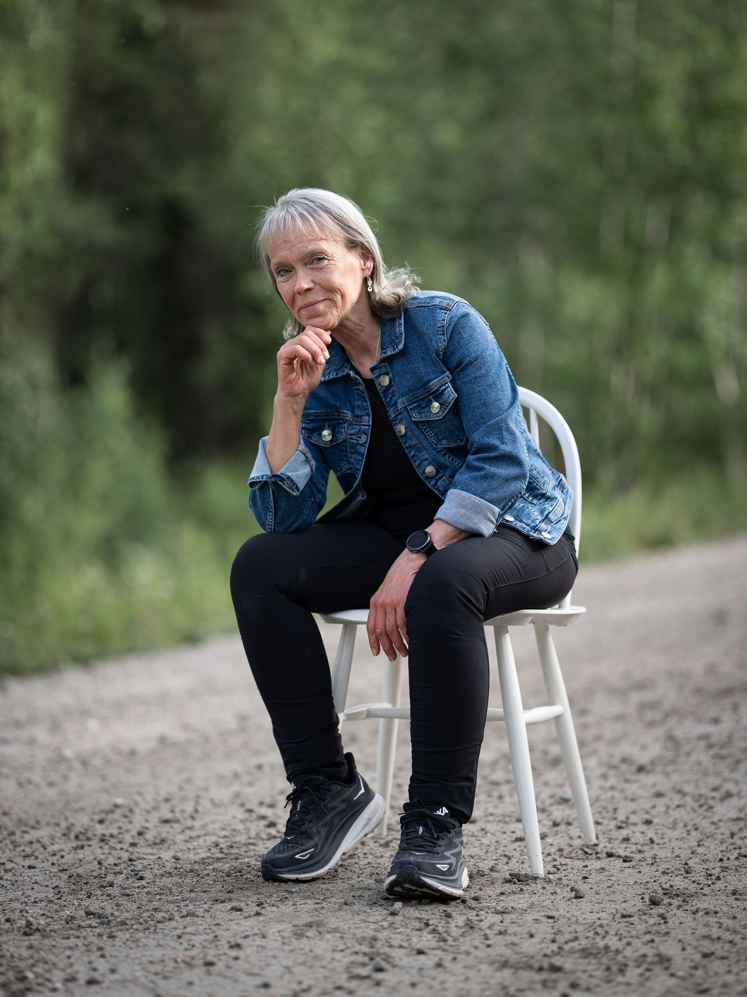 Person in denim jacket and black pants sits thoughtfully on white chair along a dirt path in nature setting.
