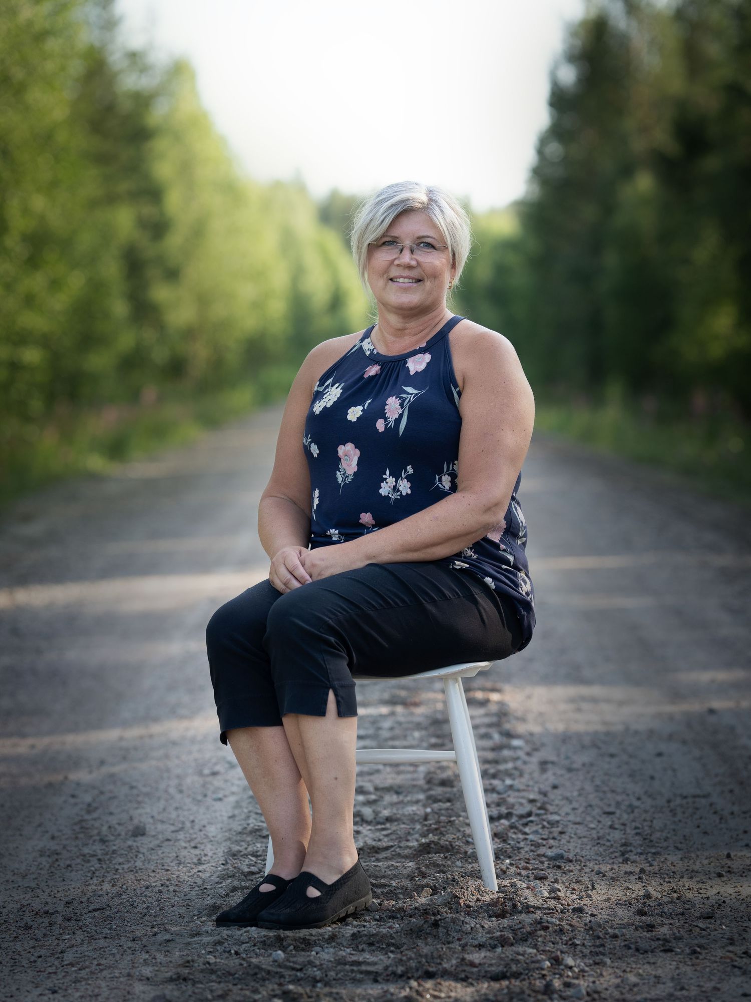 A person in a navy floral dress sits on a white chair along railroad tracks surrounded by green trees.