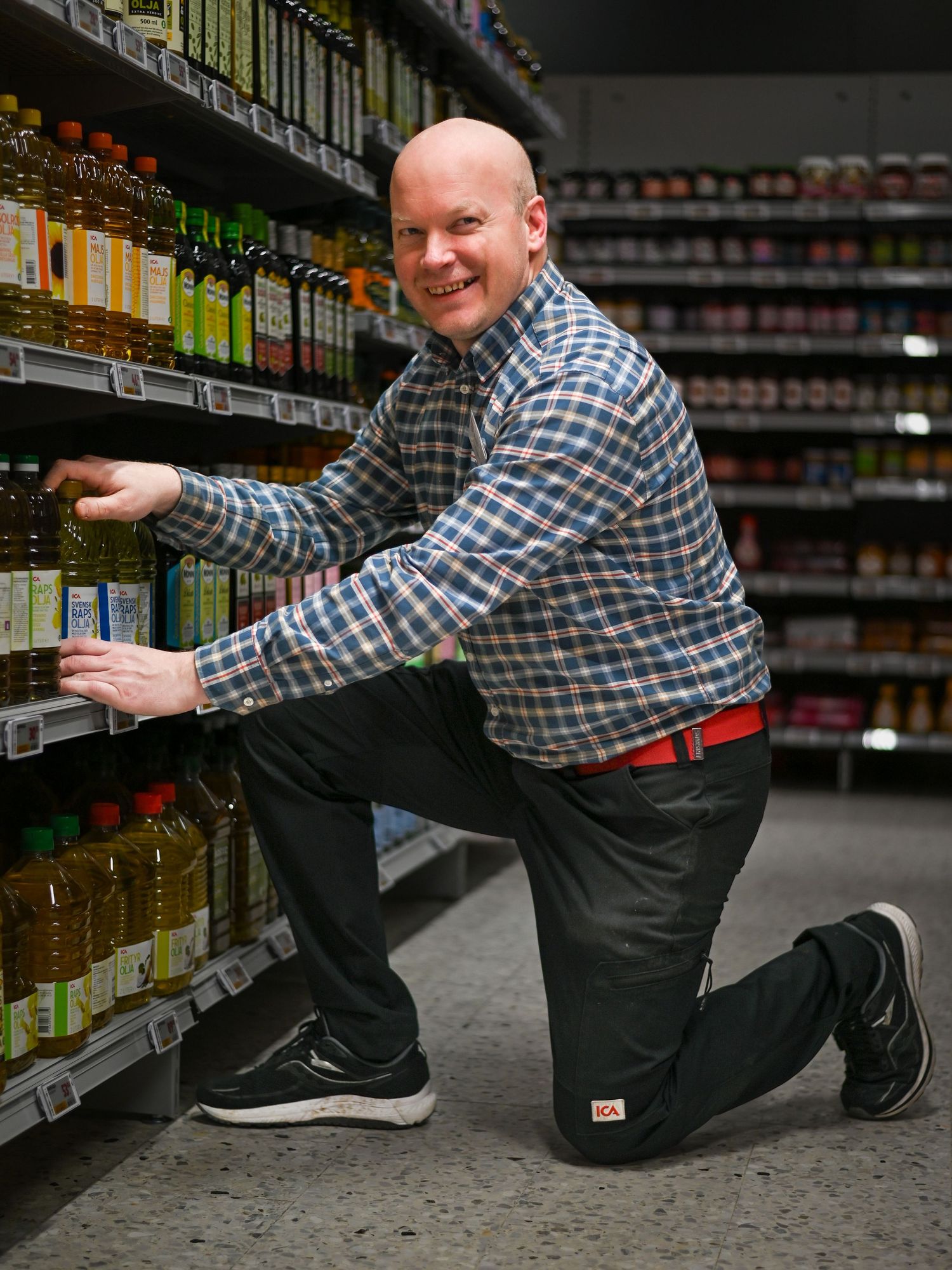 Store worker kneeling to stock shelves in grocery store aisle wearing plaid shirt and gray pants.
