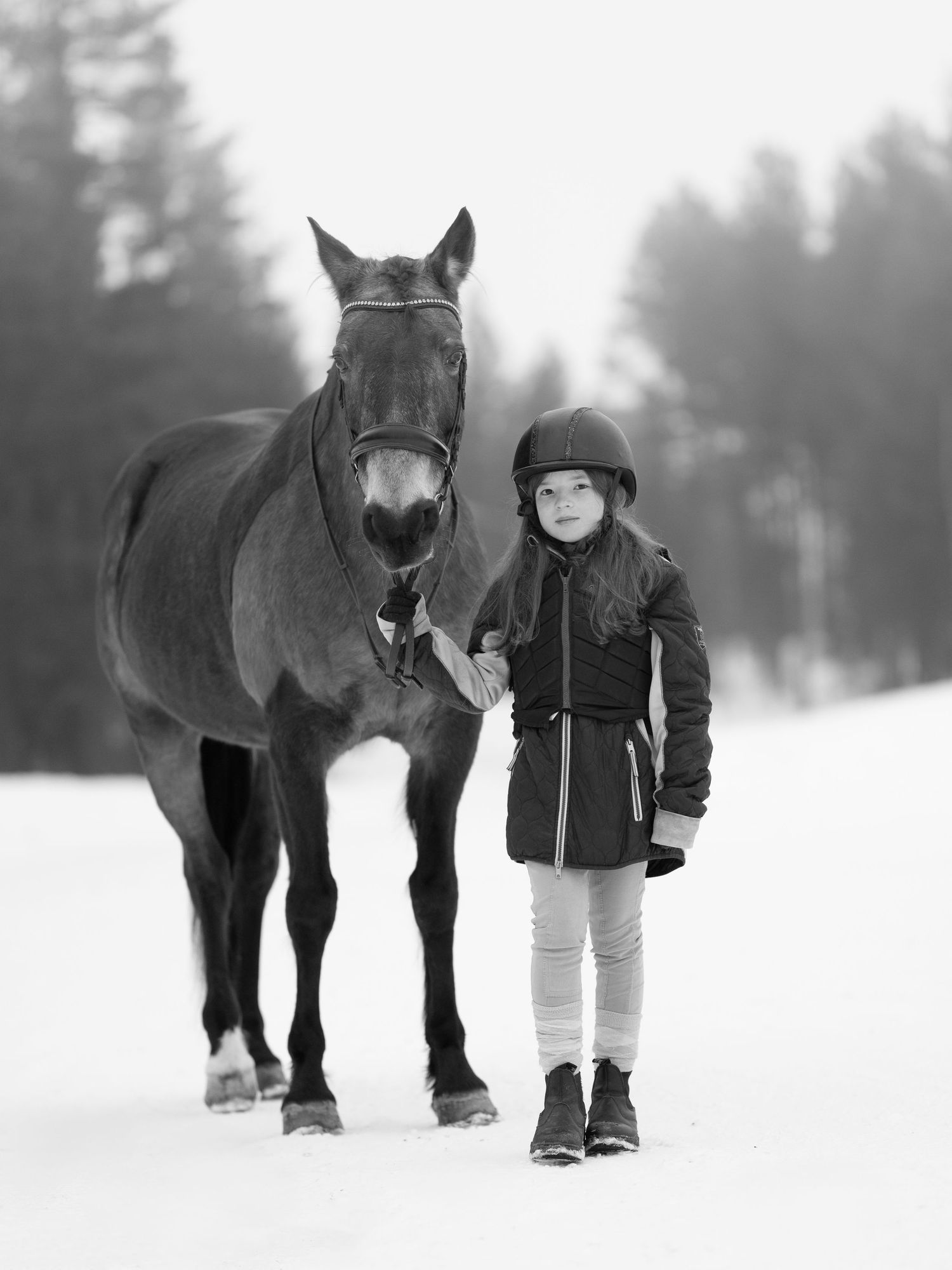 Svartvitt vinterfotografi av någon som står med en mörk häst i ett snöigt landskap.
