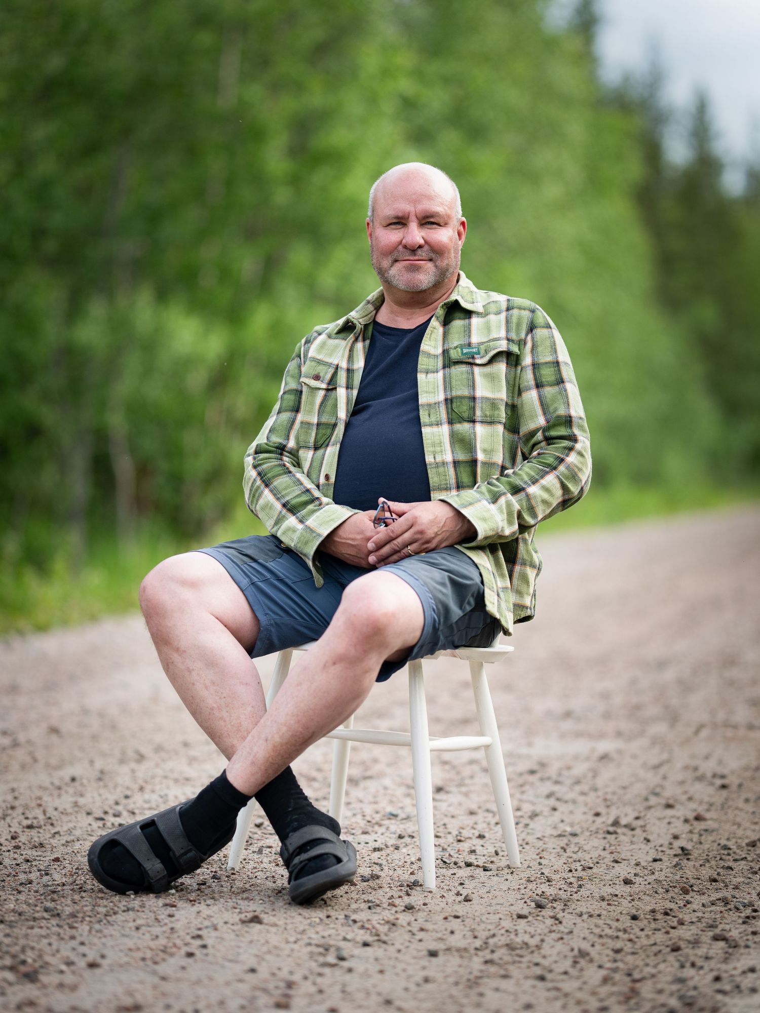 A person in a plaid shirt and shorts sits casually on a small white chair on a dirt path surrounded by green forest.