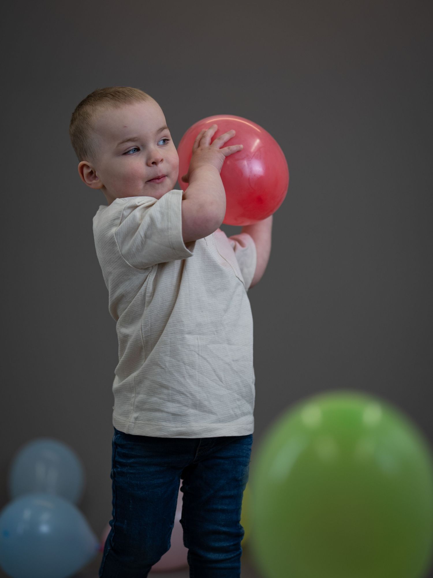 A toddler in a white shirt and jeans holds a red balloon against a dark background with other balloons visible.