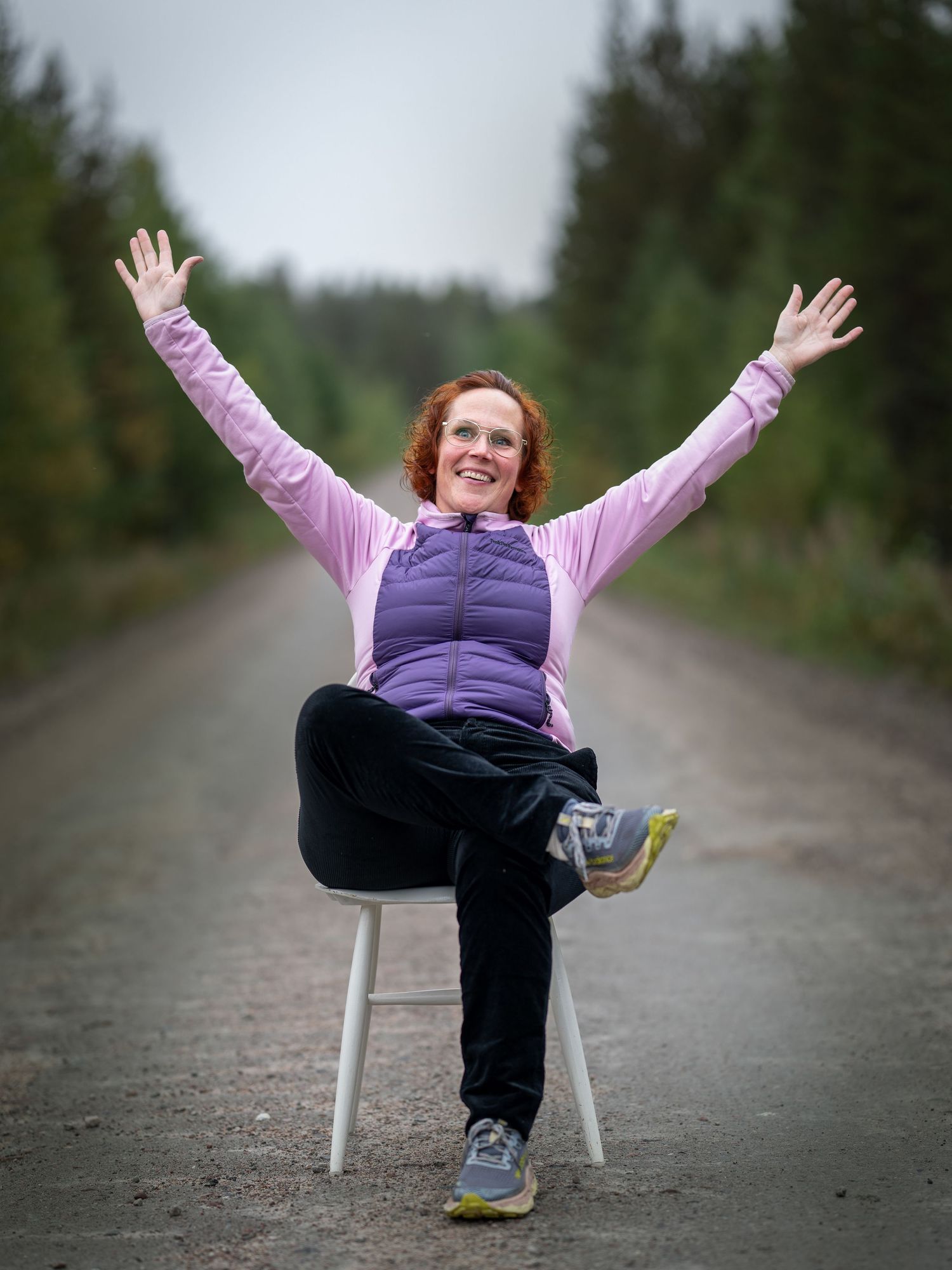 A person in pink and purple athletic wear sits on a white stool on a dirt path with arms raised joyfully.