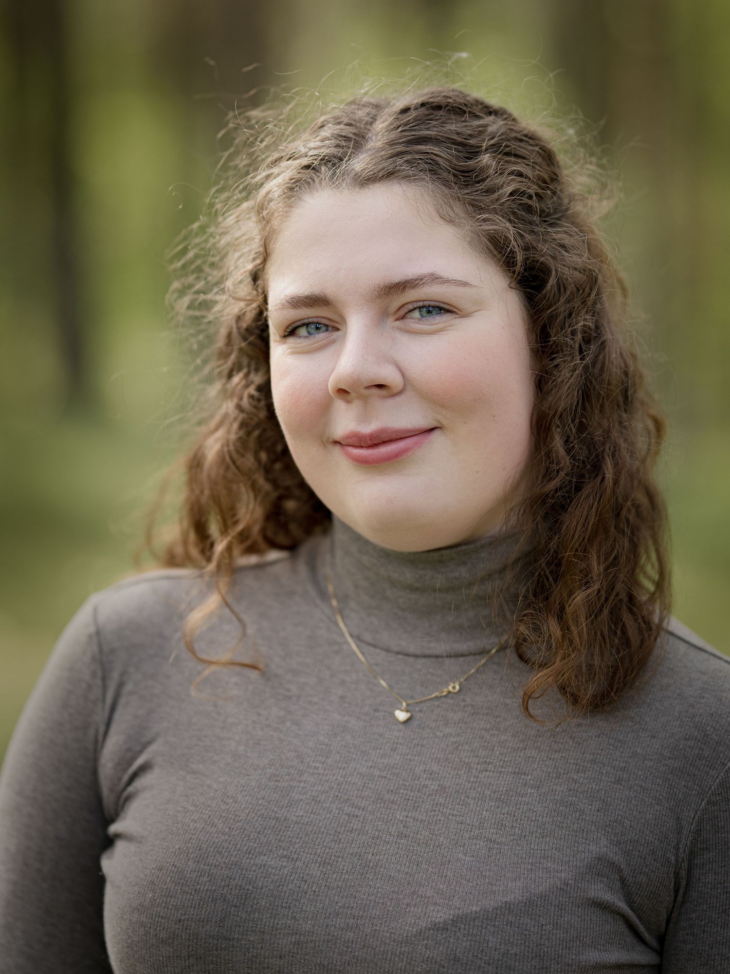 A woman with curly hair wearing a grey turtleneck smiles warmly in a natural outdoor setting.