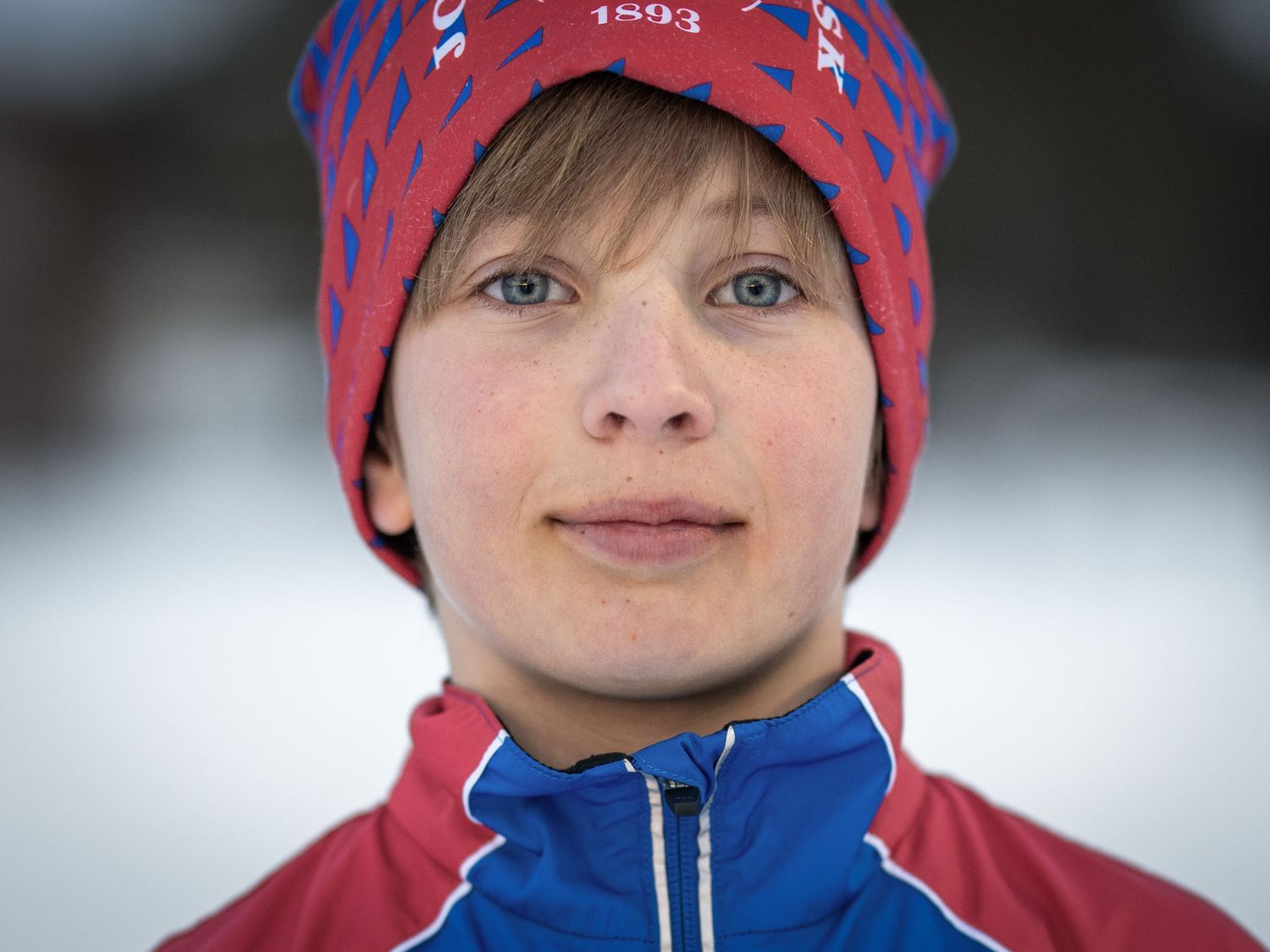 Vinteratlet bär en röd och blå mönstrad hatt och blå jacka mot snöig bakgrund.
