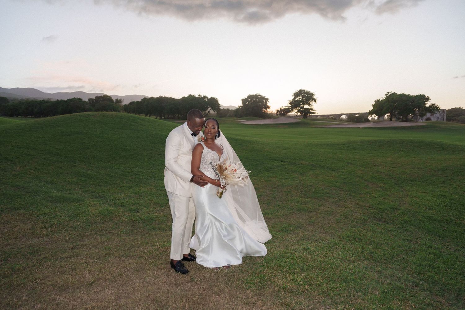 Romantic sunset portrait of newlyweds embracing on grassy hillside.