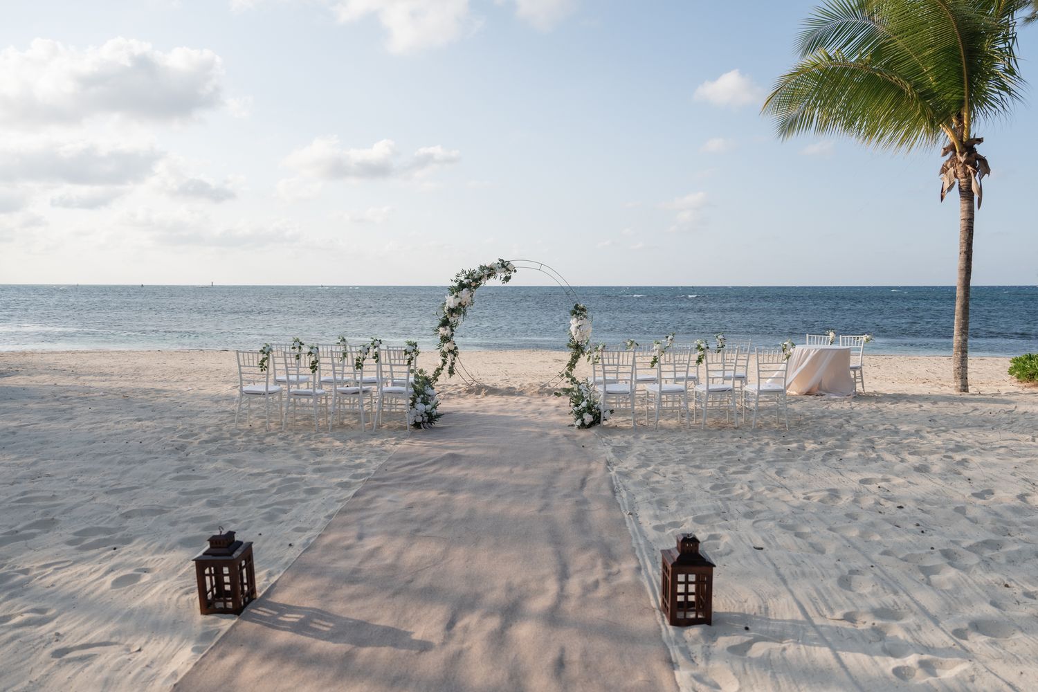 Beach wedding ceremony setup with white chairs and lanterns on sandy shore.