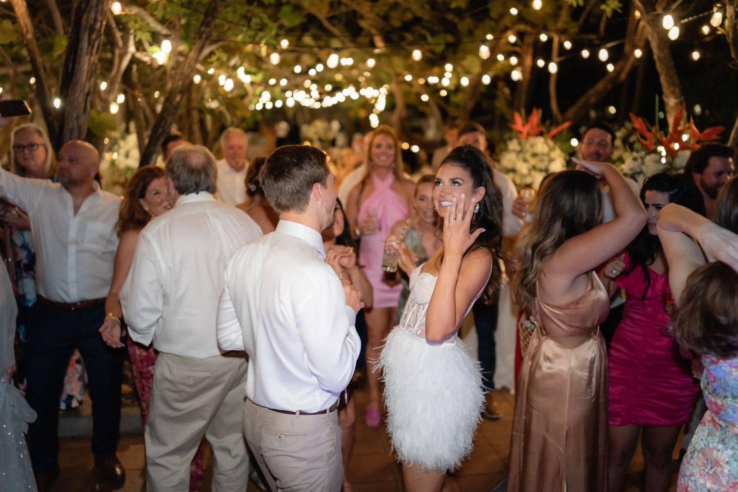 Wedding party dances under romantic string lights at outdoor evening celebration.