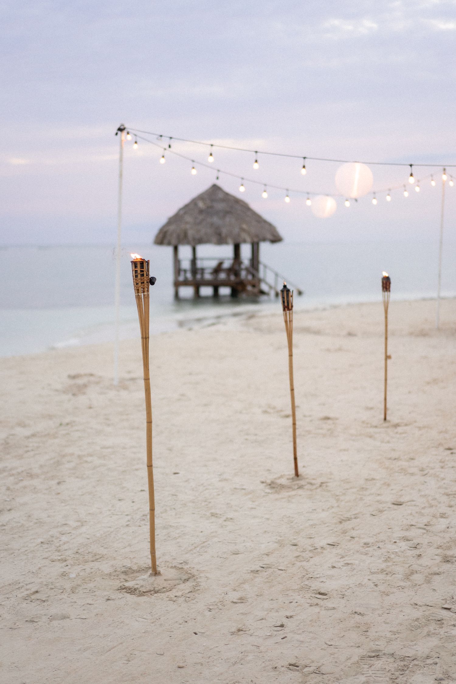 Tiki torches and string lights lead to a wooden gazebo on a beach at dusk.