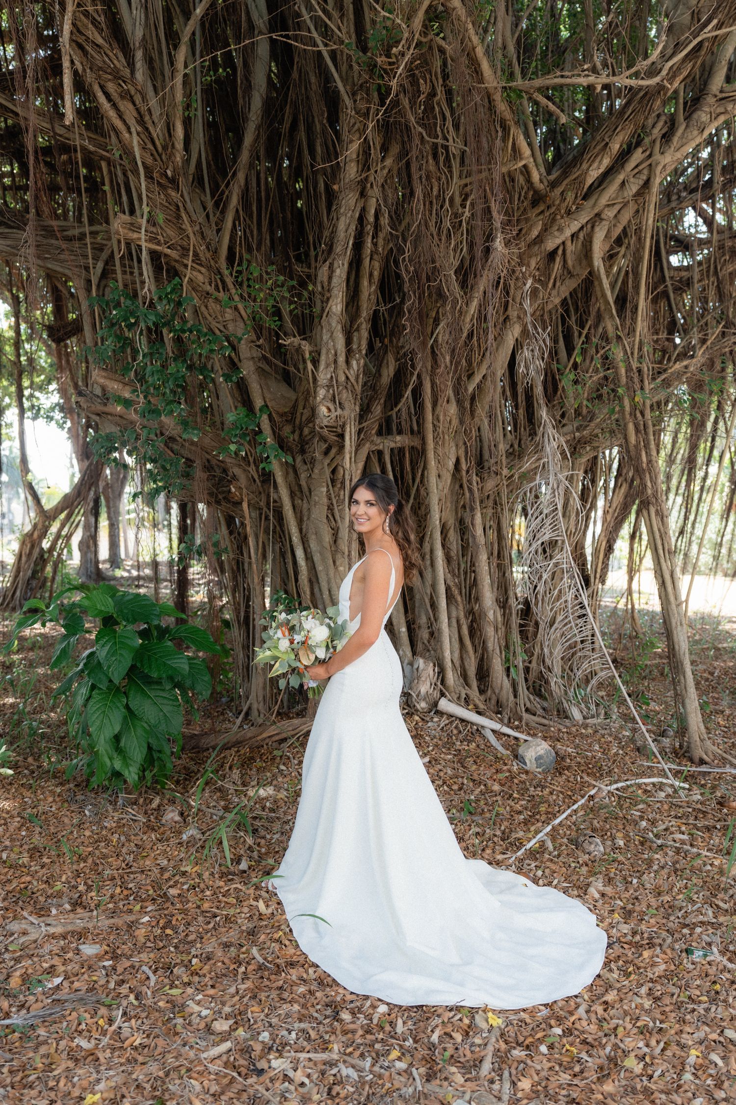 Bride poses with bouquet against dramatic banyan tree backdrop in outdoor setting.
