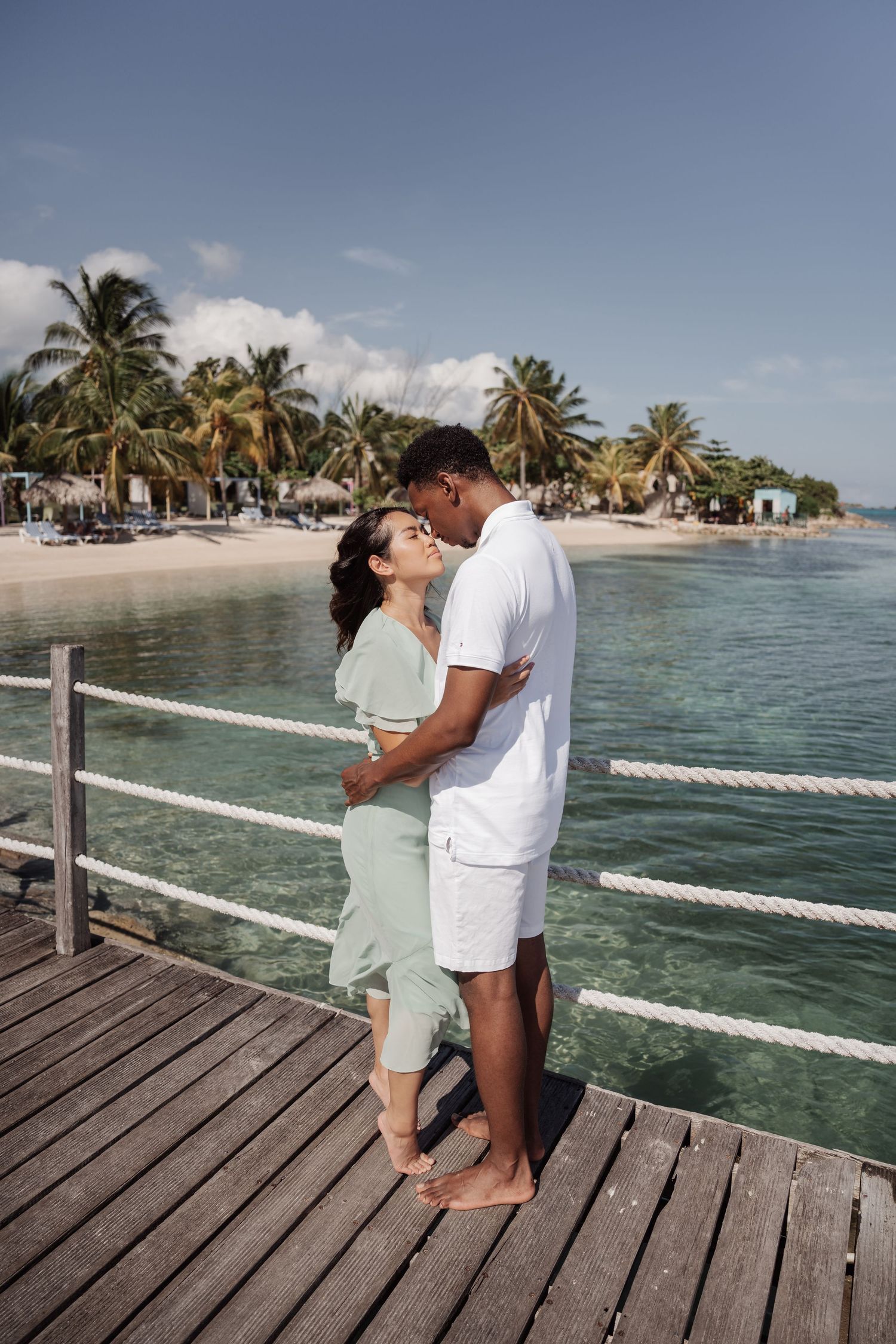 A couple shares an intimate moment on a wooden deck overlooking turquoise waters and palm trees.