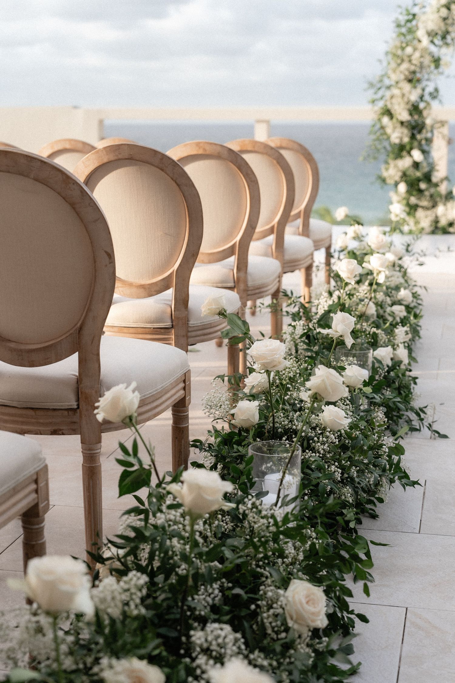 Elegant wedding ceremony chairs lined with white roses and greenery along a waterfront setting.