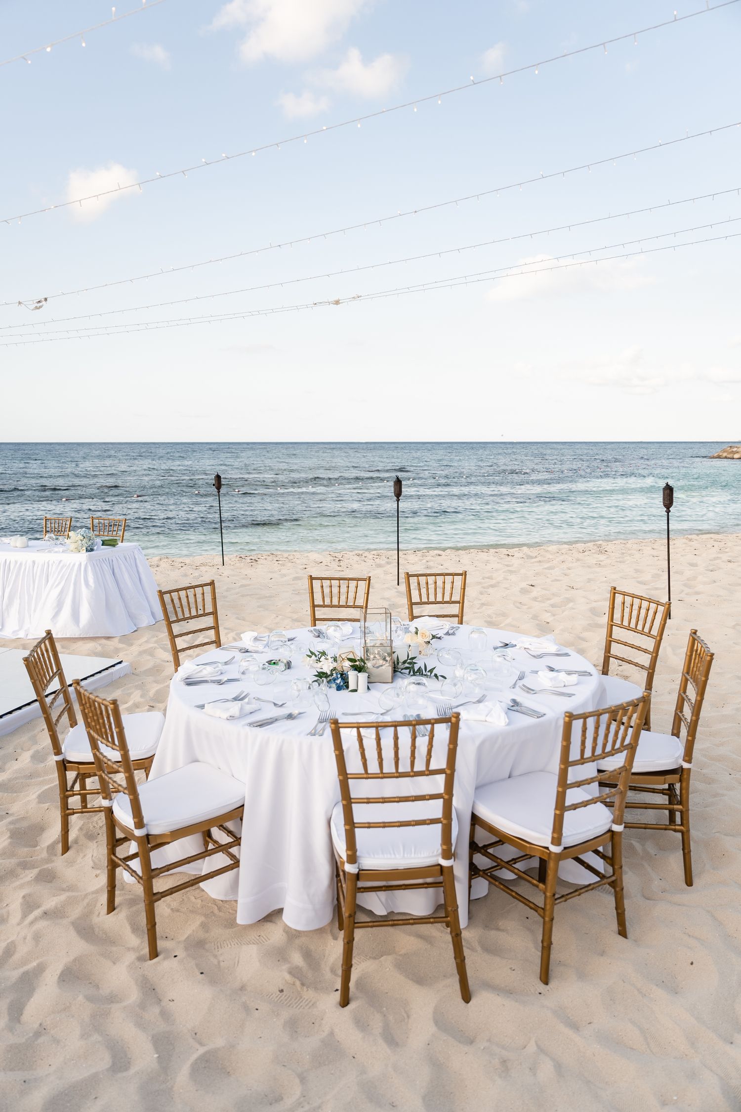 Round dining table set with white linens and gold chairs on beach for intimate reception.