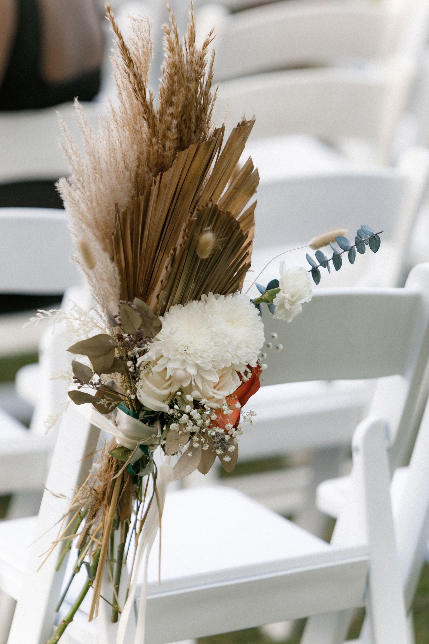 A rustic wedding aisle decoration featuring dried pampas grass, white flowers and eucalyptus on a white chair.