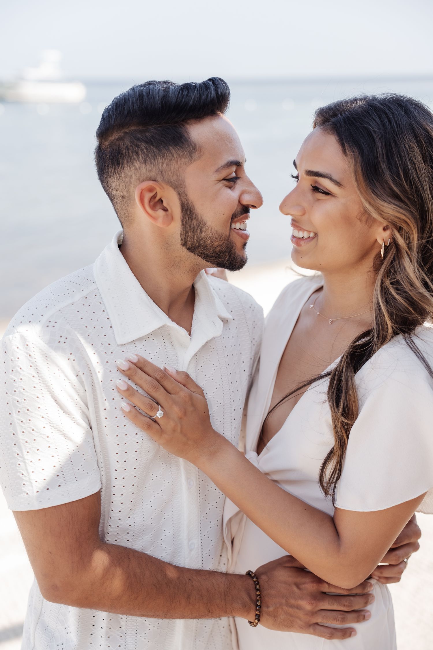 Couple in white outfits embracing and smiling at each other on a sunny beach.