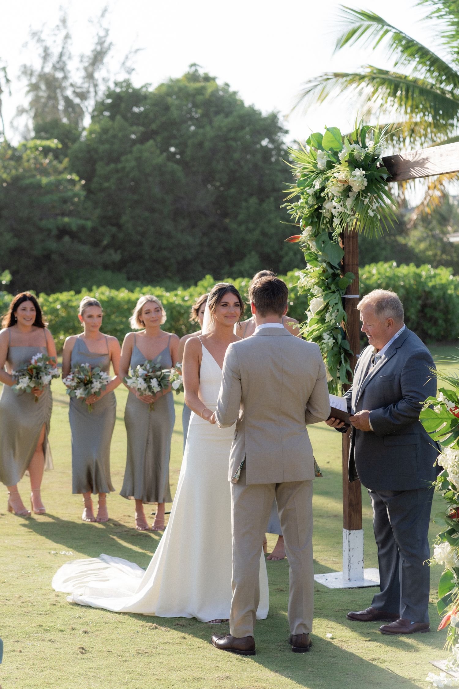 A wedding ceremony takes place on a lush lawn with tropical foliage and bridesmaids in grey dresses.
