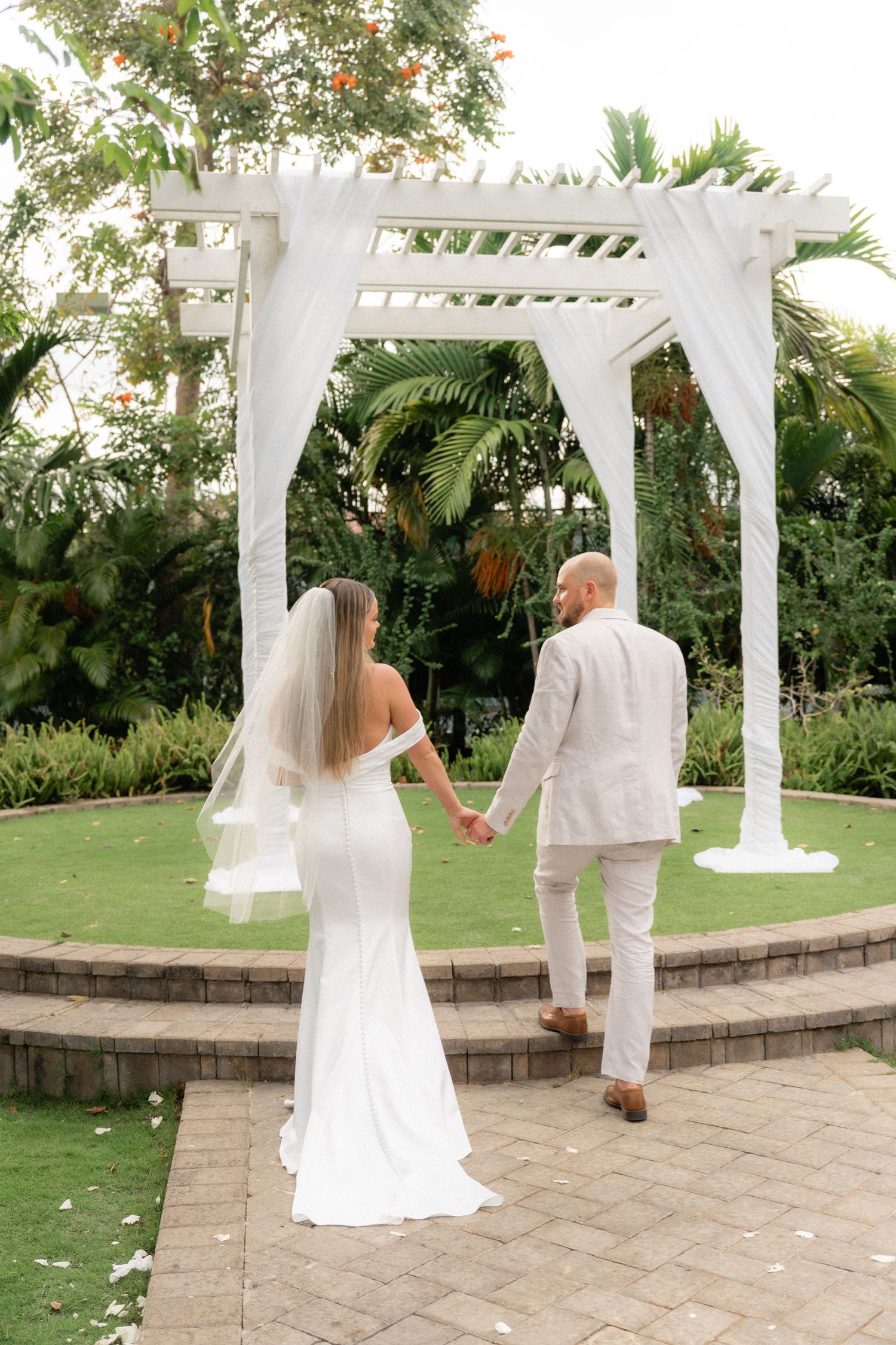 Newlyweds walking hand in hand towards white pergola in tropical garden setting.