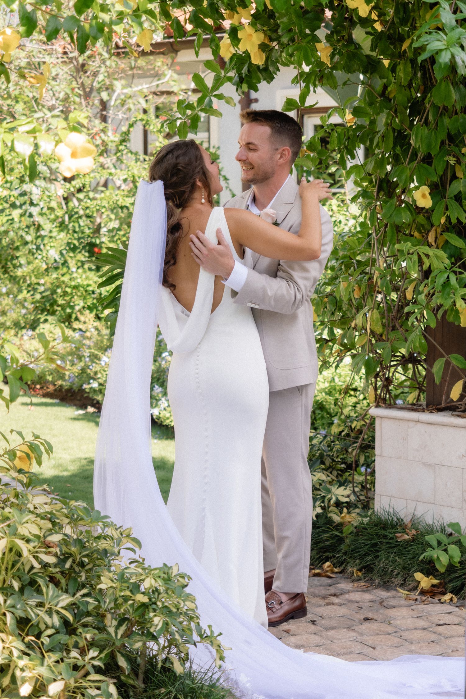 Wedding couple embraces under garden pergola with cascading greenery and yellow flowers.