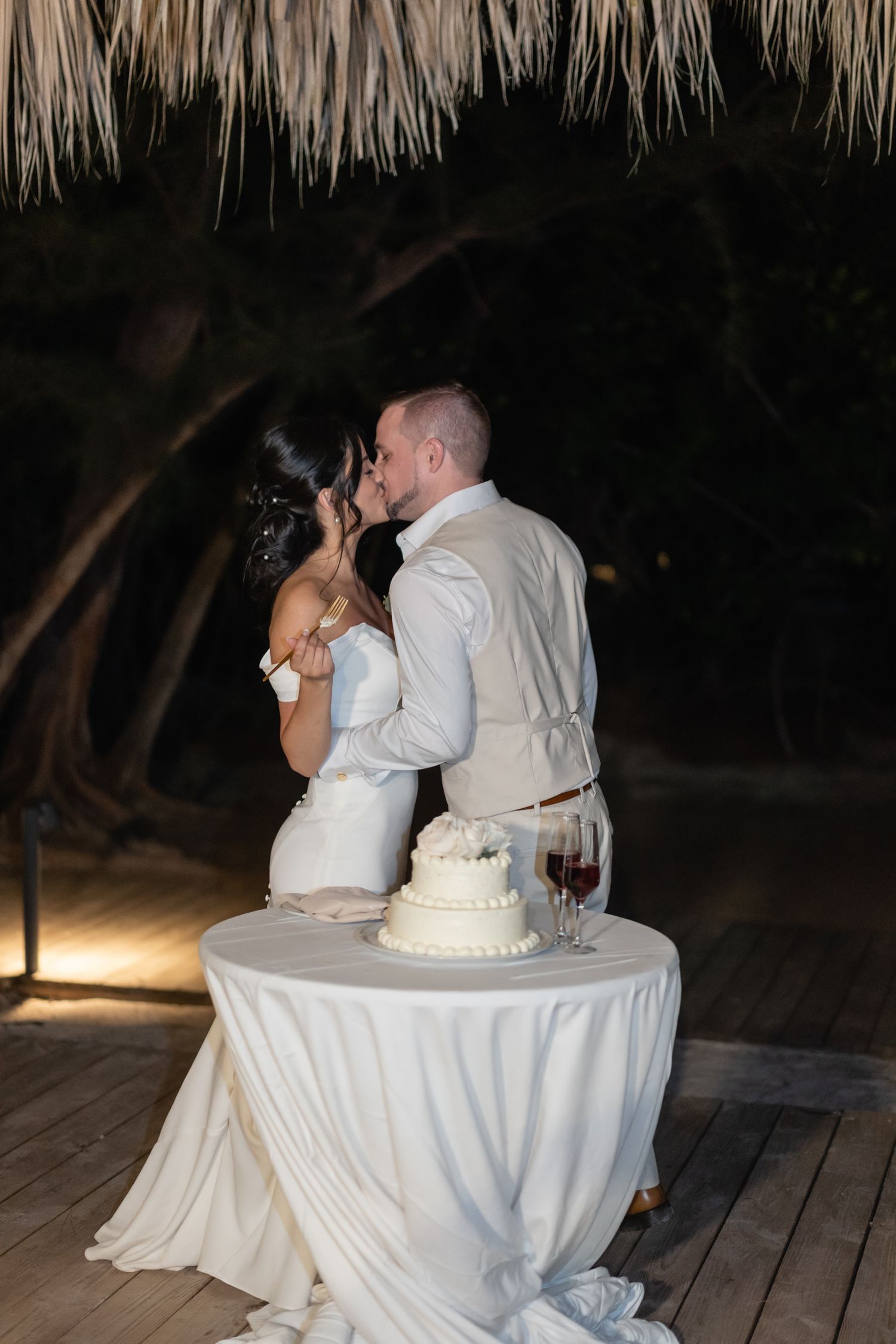 Wedding cake cutting moment under thatched roof on tropical beach deck at night.