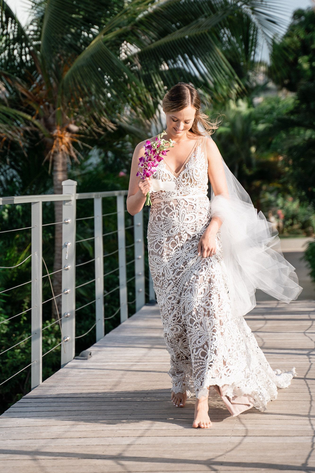 A bride in a delicate lace dress walks barefoot on a wooden walkway surrounded by palm trees.