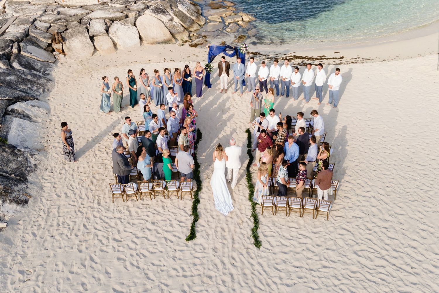 Birds-eye view of beach wedding ceremony with guests seated in organized rows on white sand.