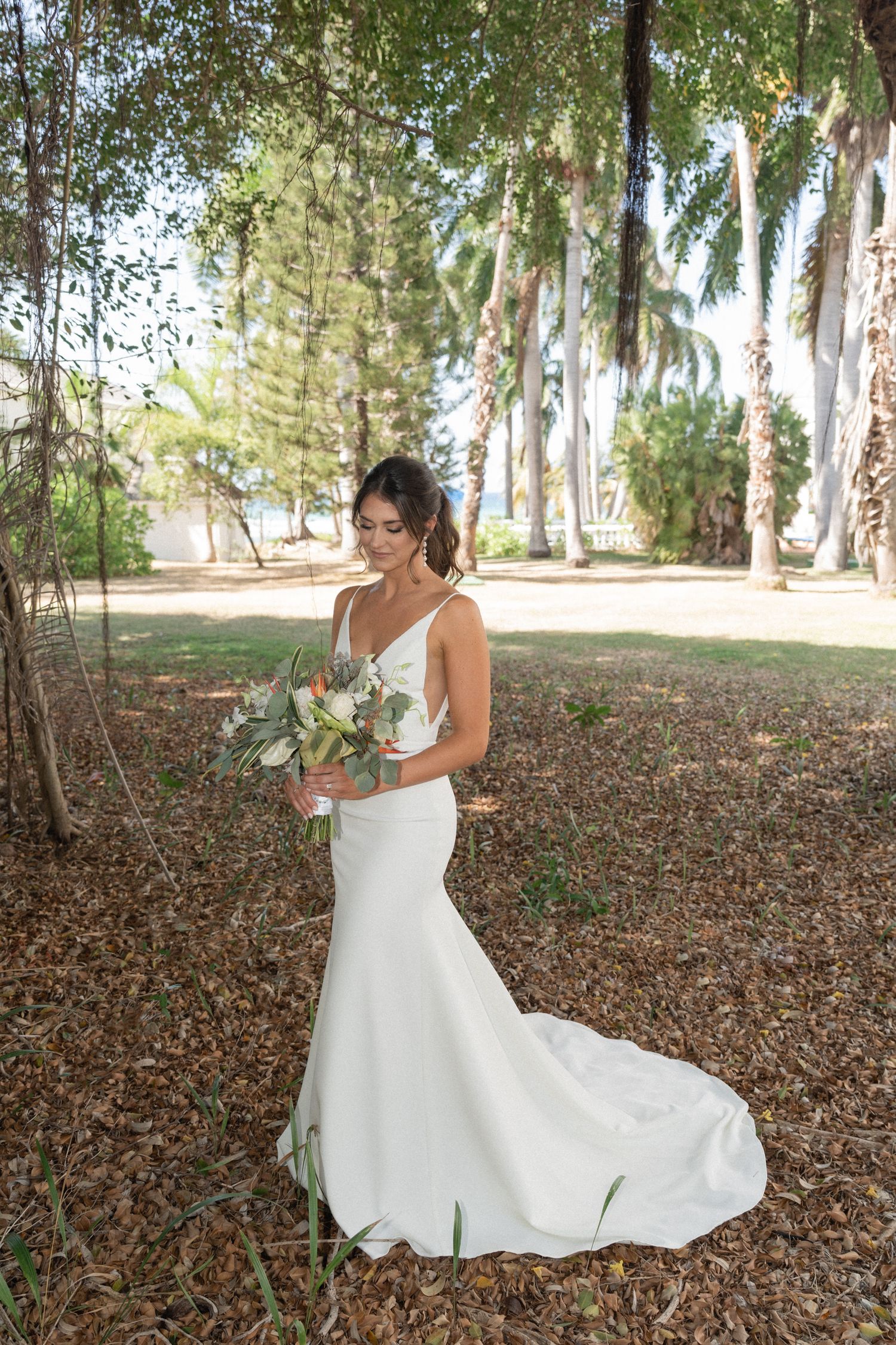 Bride in fitted white gown with train poses among palm trees and fallen leaves.