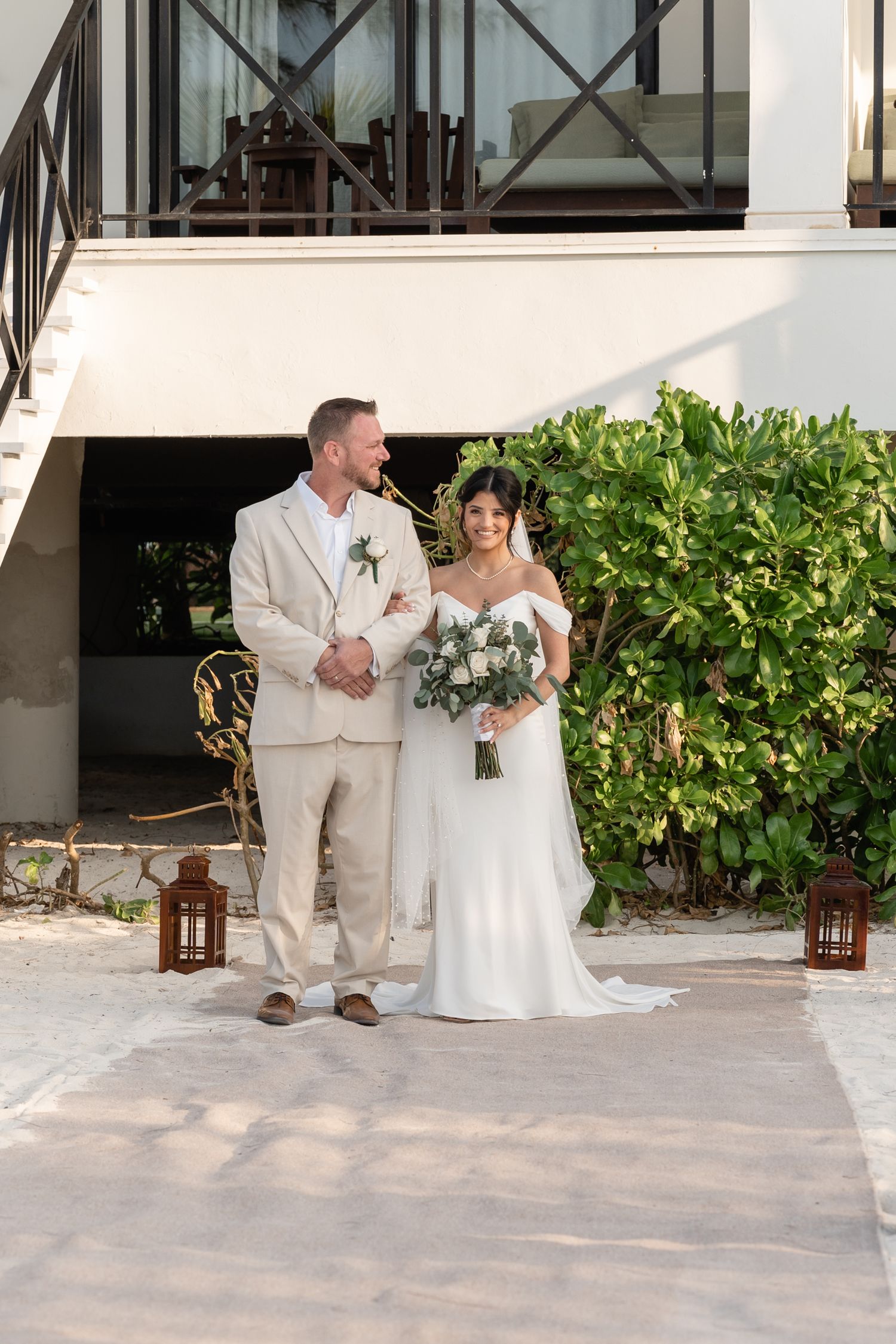 A wedding couple poses together on a paved pathway adorned with lanterns and tropical foliage.