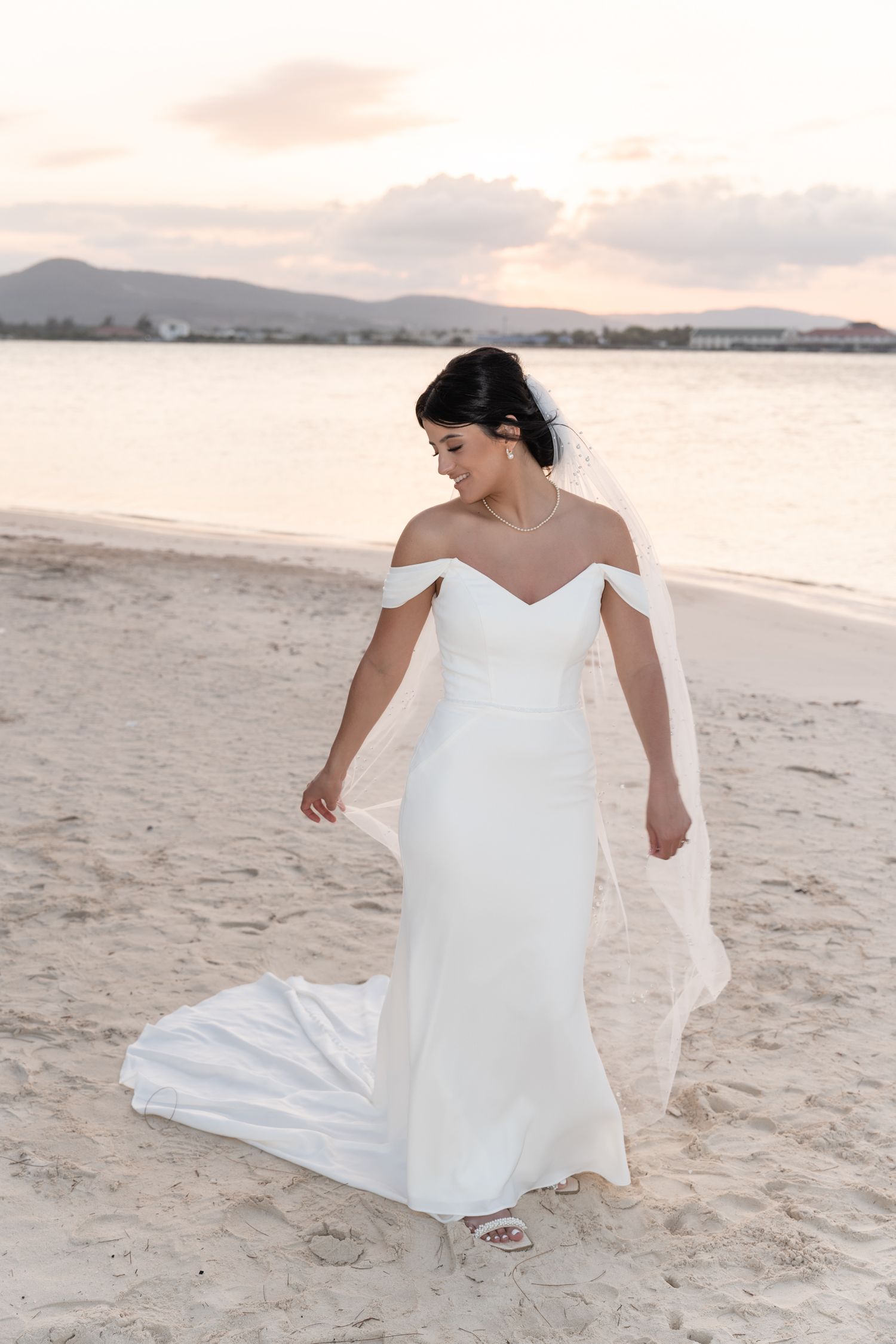Elegant bride in off-shoulder white dress walking on beach at sunset with mountains in background.