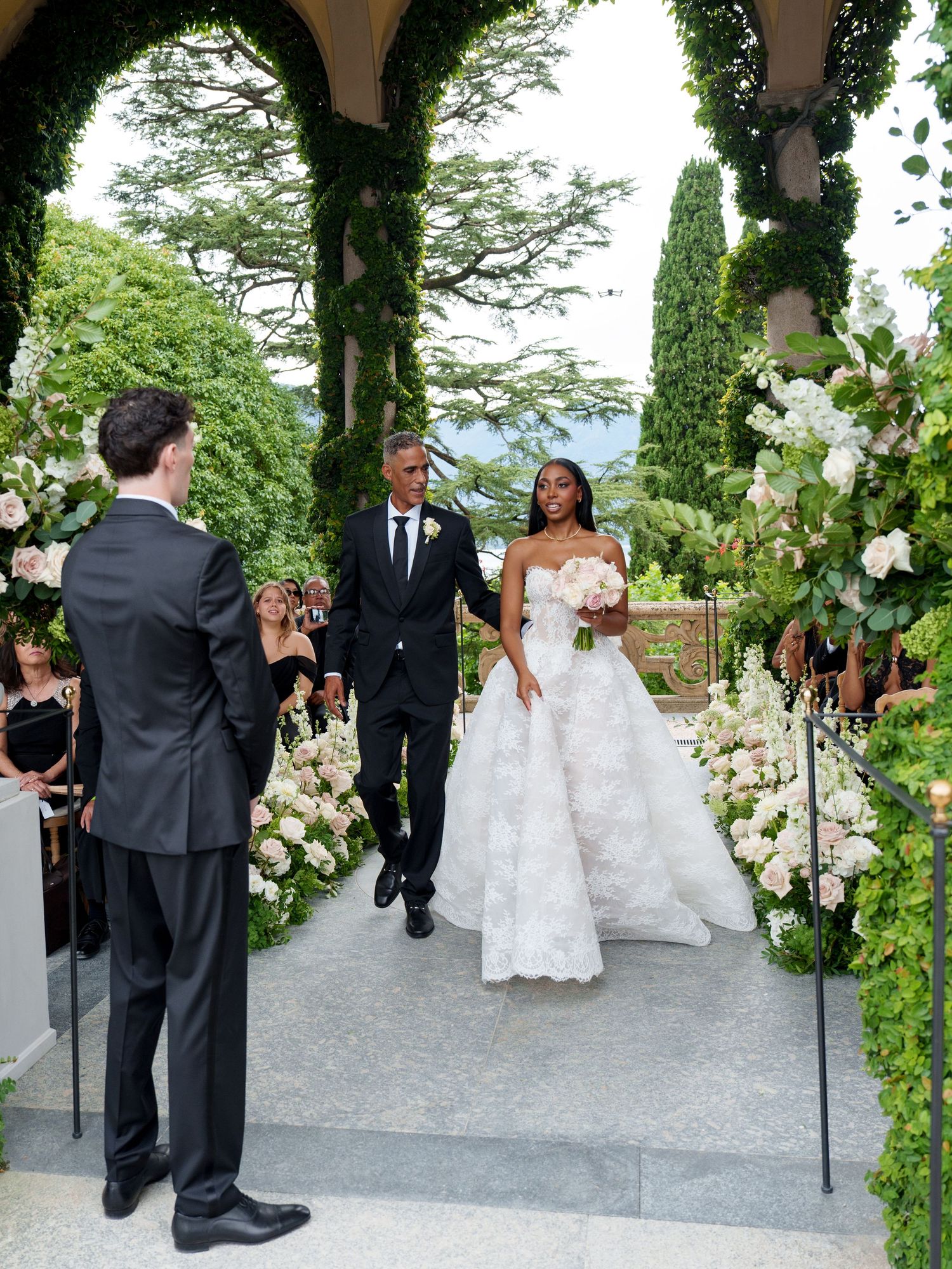 Wedding party processes through a lush garden path lined with cypress trees and white floral arrangements.