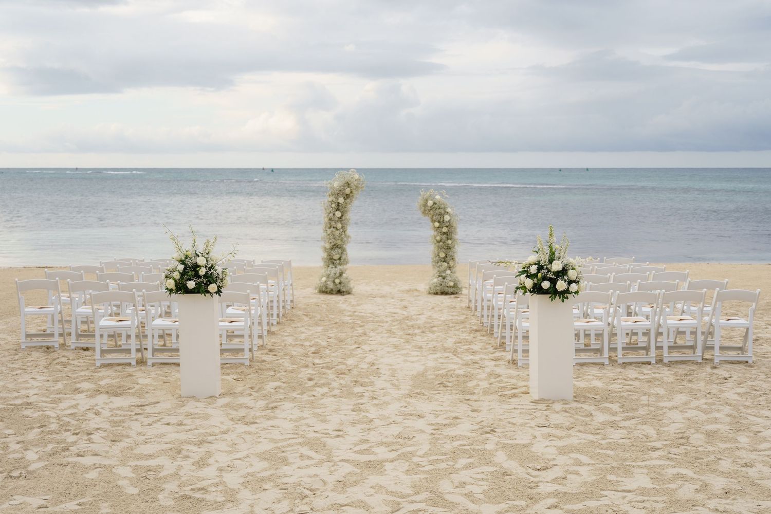A romantic beach wedding setup with white chairs and floral archway on the sandy shore overlooking the ocean.