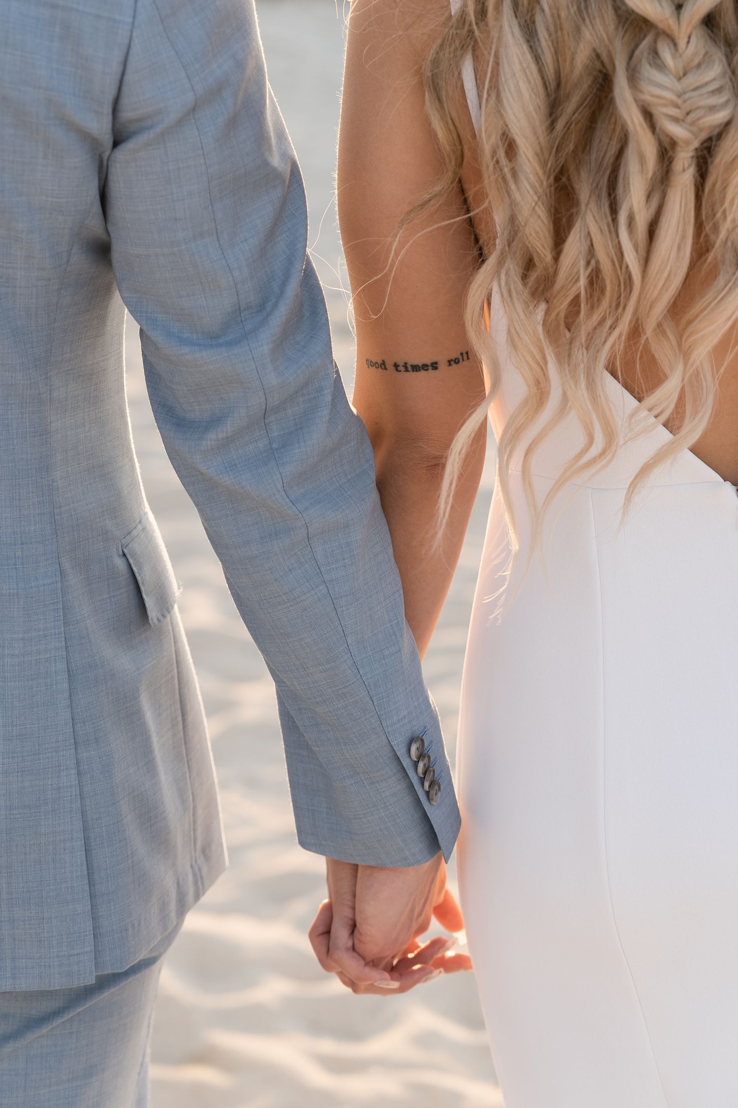 Close-up of a couple holding hands on the beach, showing elegant wedding attire and a small tattoo.