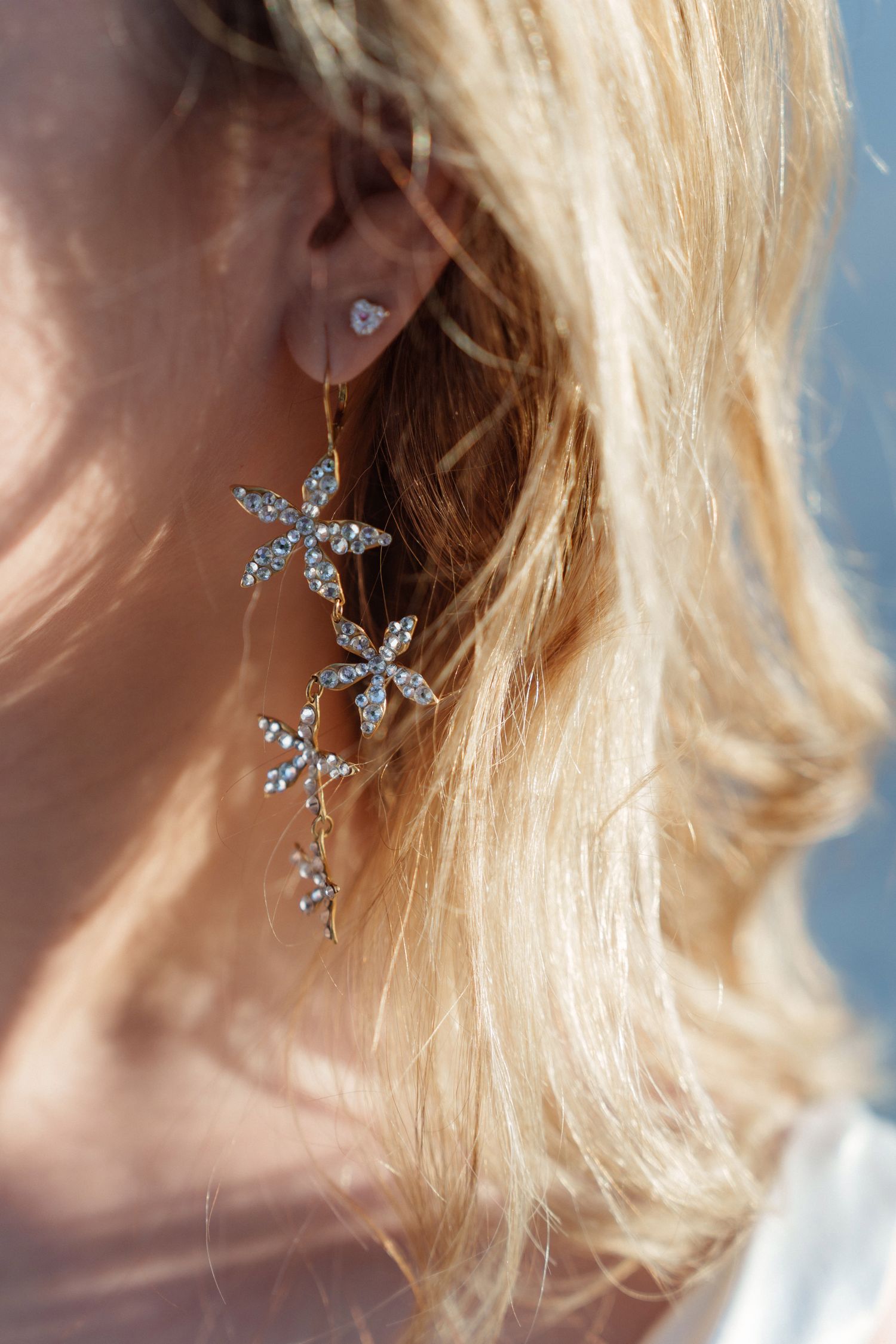Crystal flower earring dangling against blonde hair with ocean background.