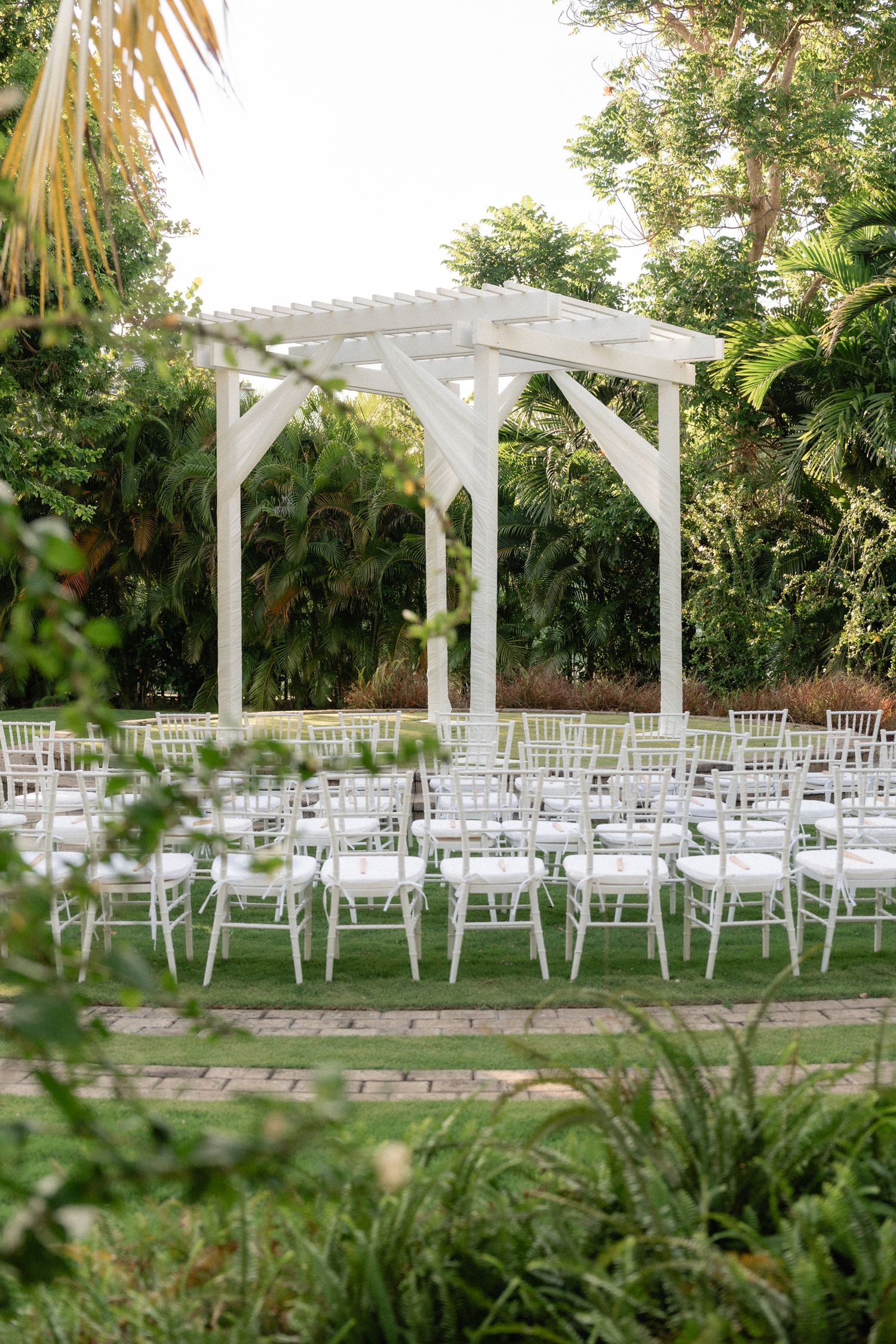 White garden pergola set up for outdoor wedding with rows of white chairs on stone patio.