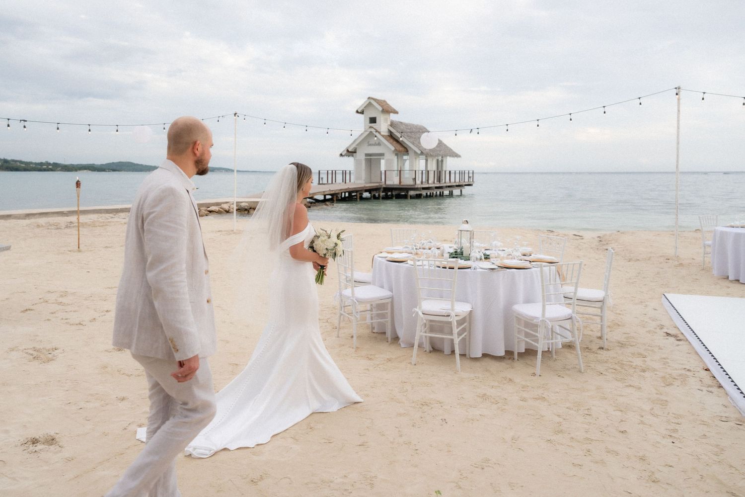 Wedding reception setup on beach with pier gazebo and white dining tables.
