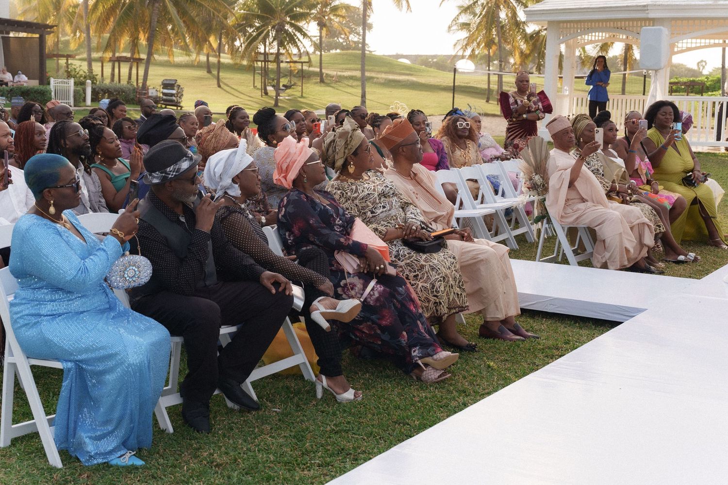 Guests seated on white chairs at an outdoor wedding with palm trees and ocean views.