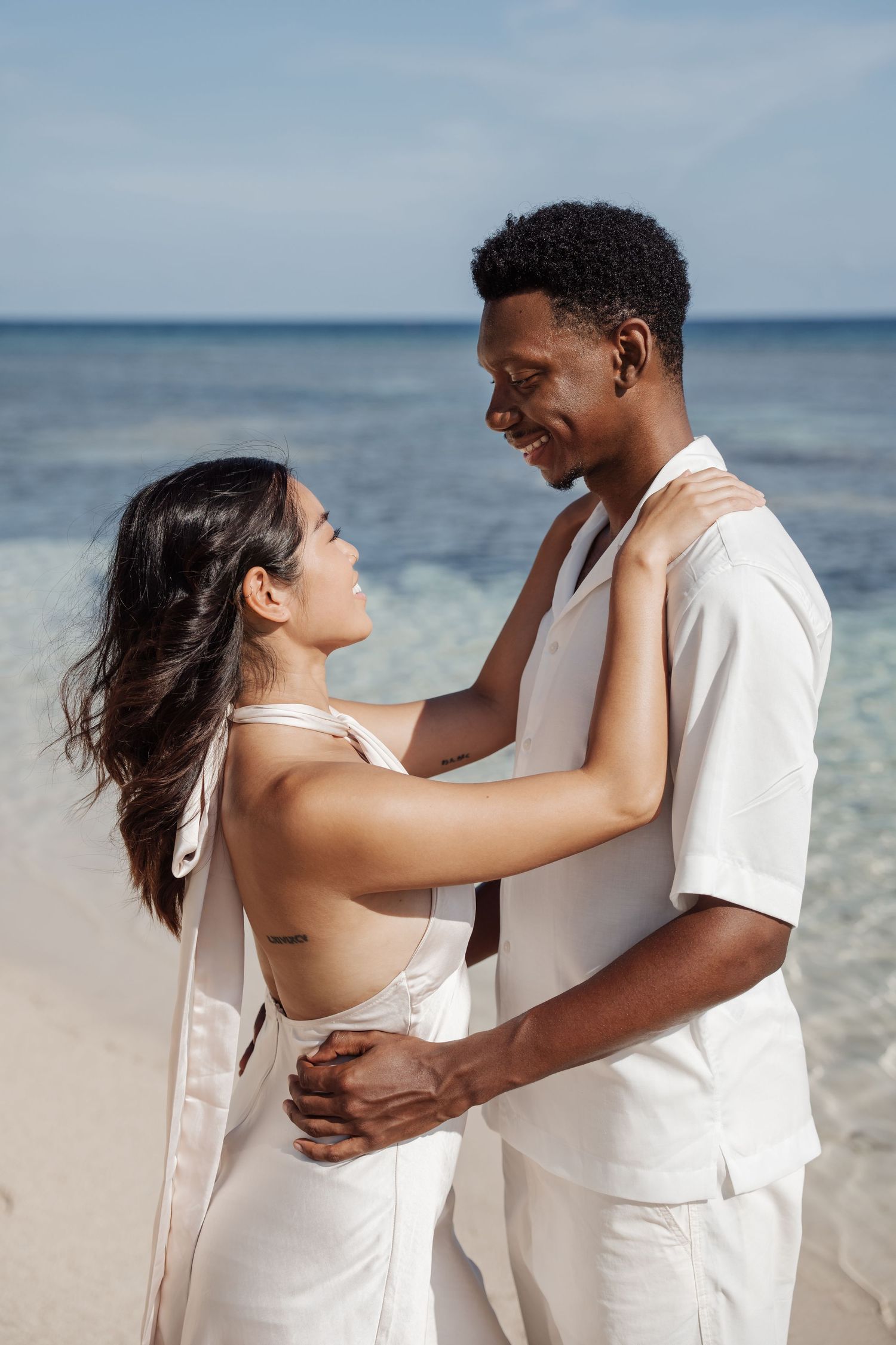A couple in white outfits embraces on a sunny beach with crystal clear waters behind them.