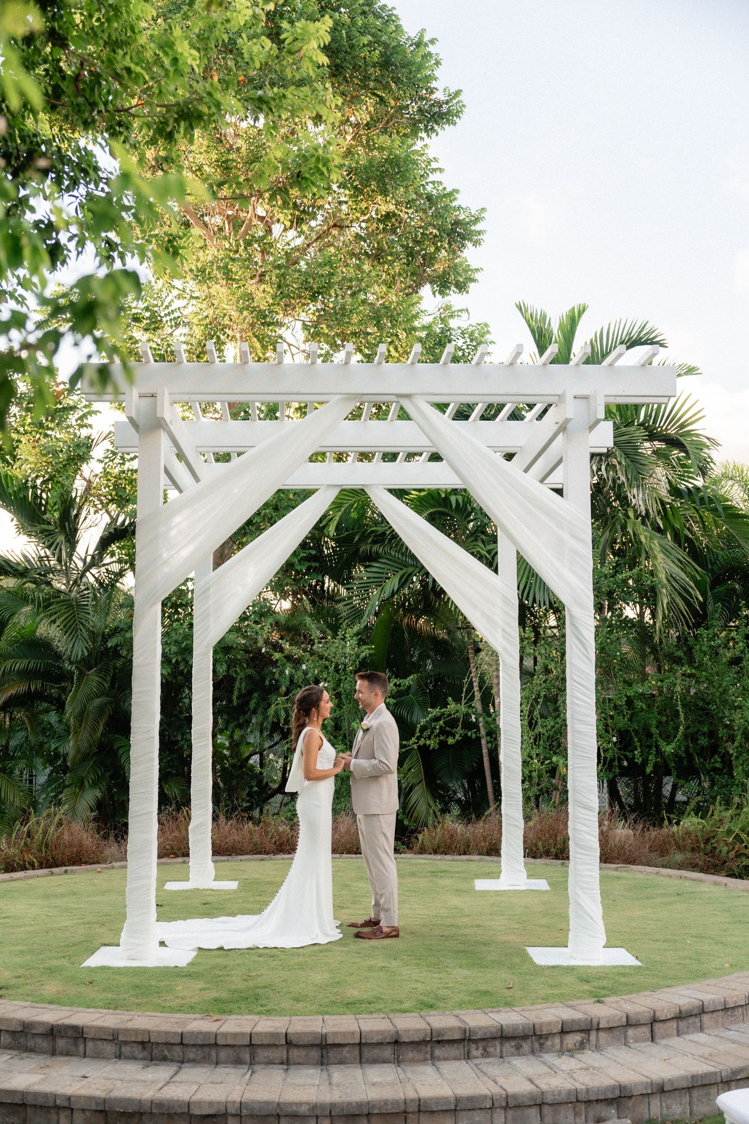 Couple stands face-to-face under white wooden pergola during intimate wedding ceremony.