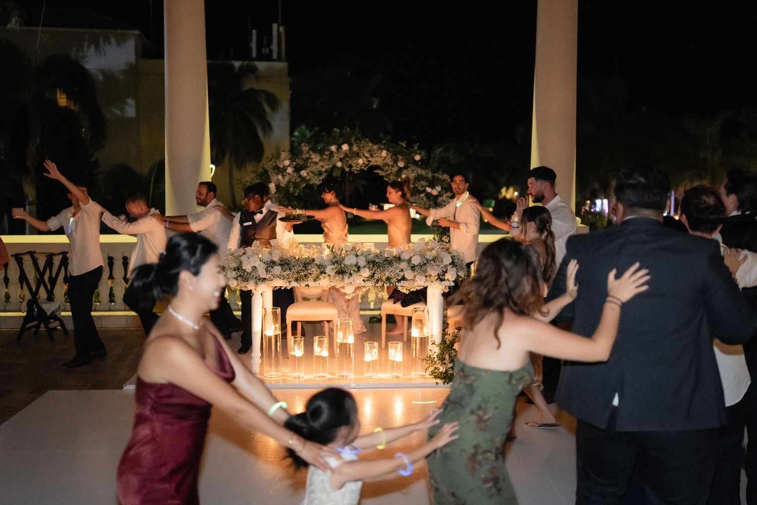 Wedding guests dance in a circle at an evening reception with candlelit tables in background.