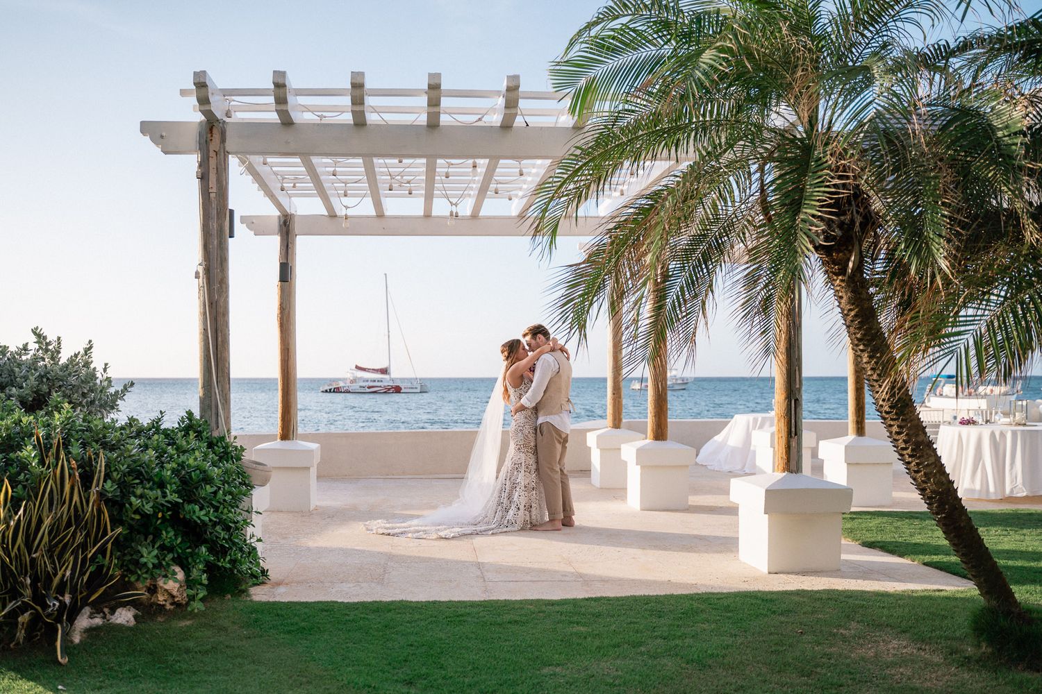 A beachside ceremony setup with wooden pergola, palm trees, and ocean view in the background.