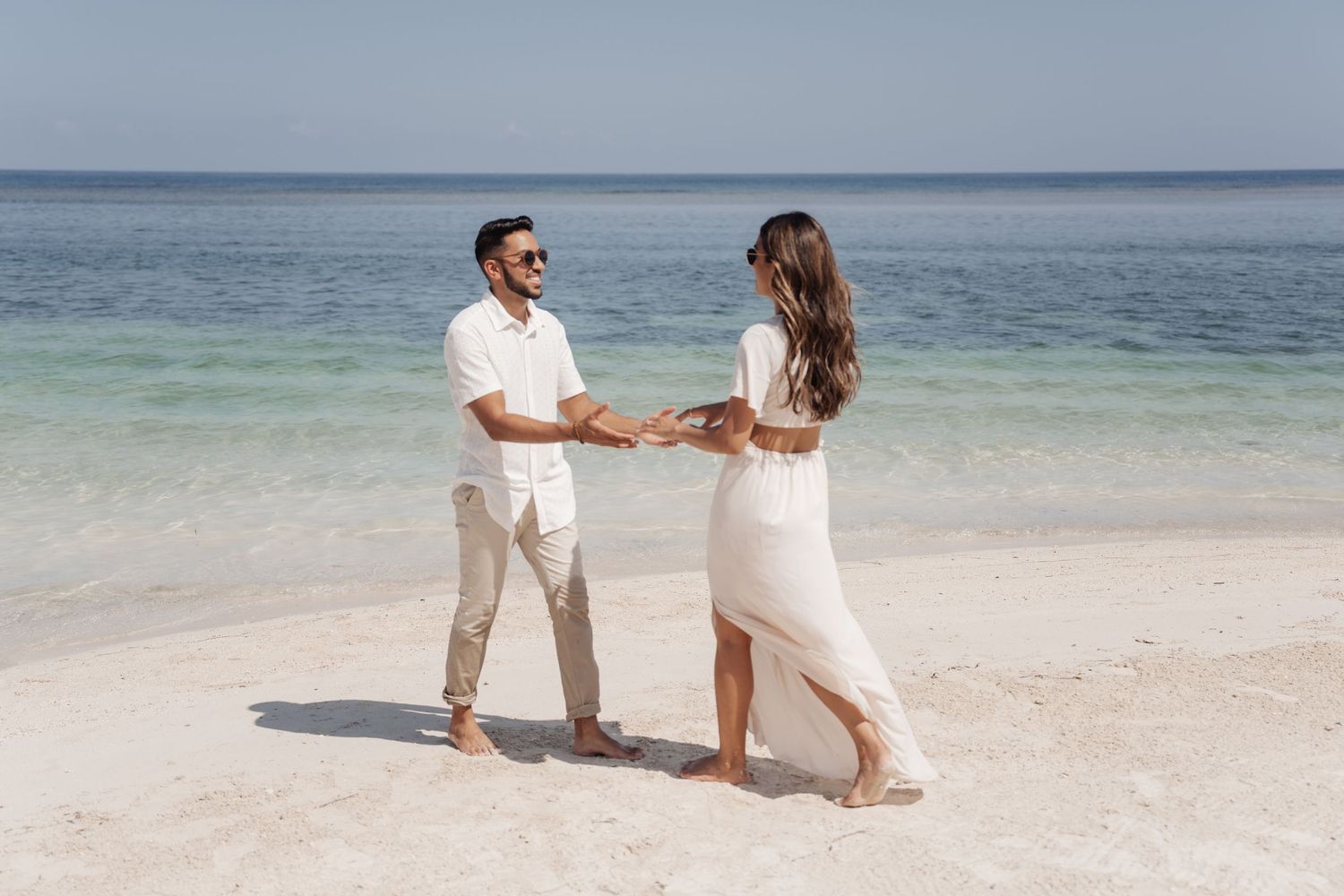 Couple in white holding hands while dancing on a pristine beach with turquoise waters.