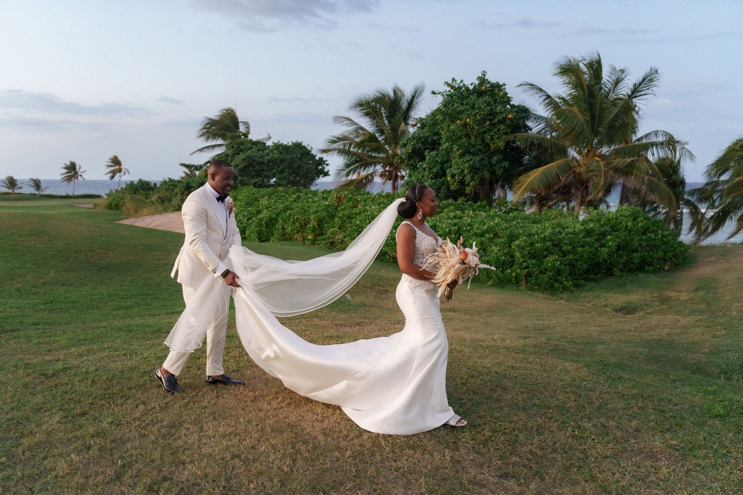 Couple poses with flowing wedding dress train on green oceanfront lawn.