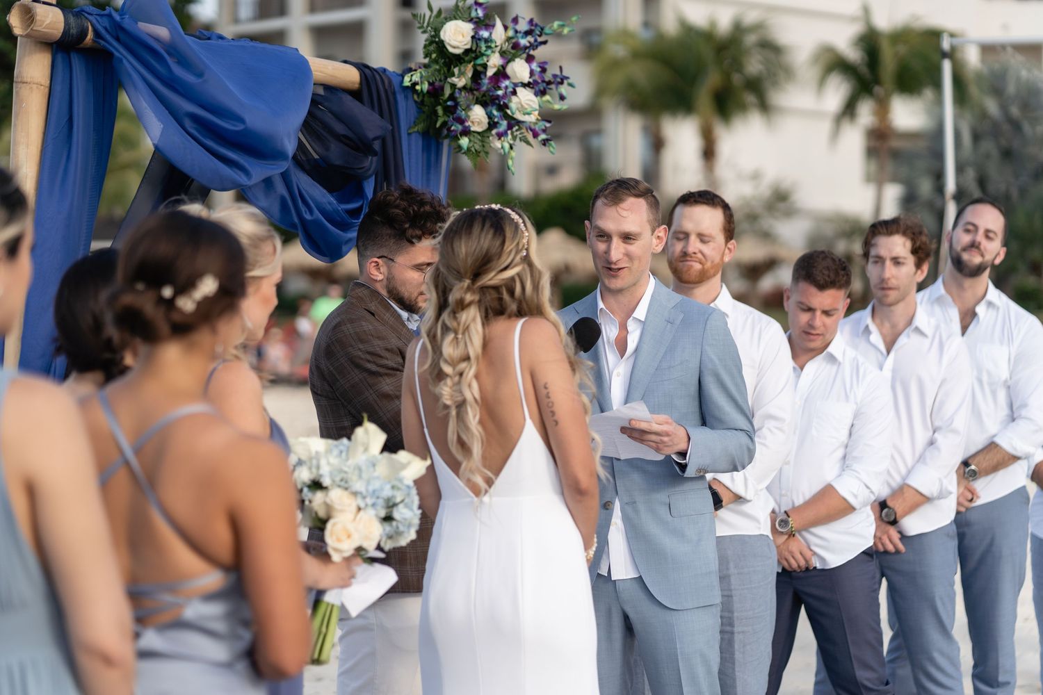 Wedding ceremony on beach with bridal party in blues and greys under draped arch.