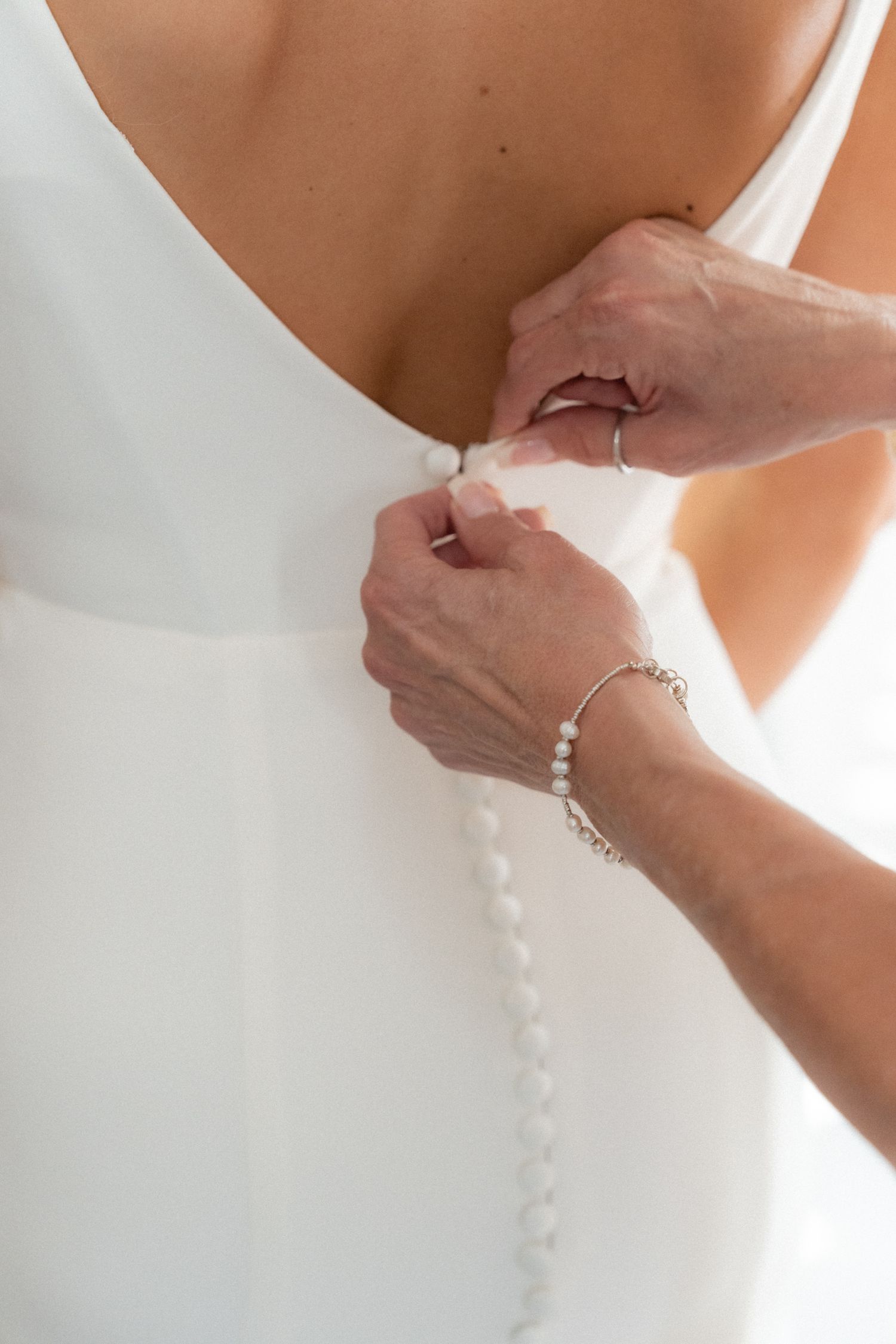 Close-up detail of hands buttoning the back of an elegant white wedding dress.