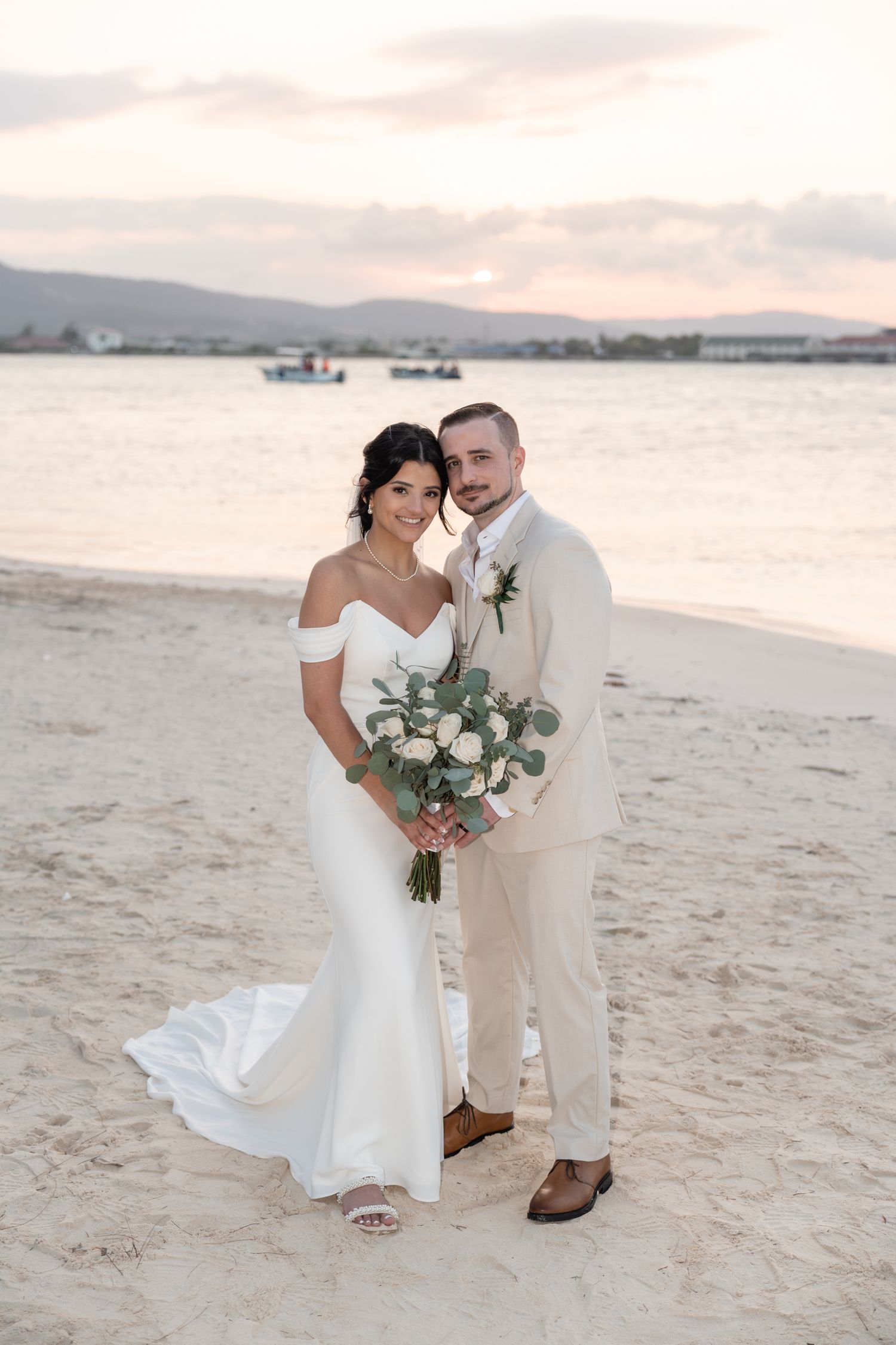 Romantic beach portrait at sunset with mountains and boats in background.