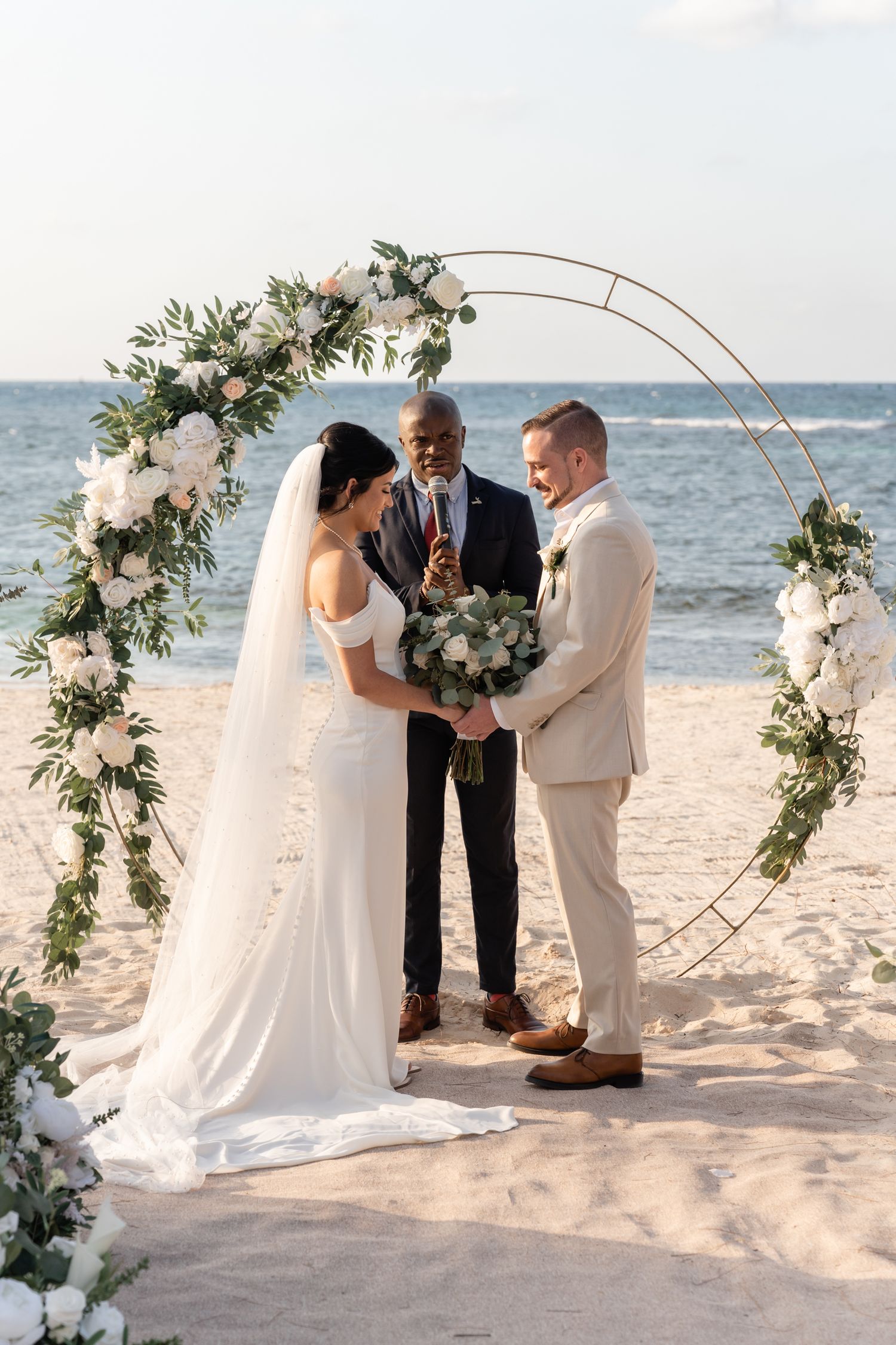 Beach wedding ceremony under a circular floral arch with ocean backdrop.