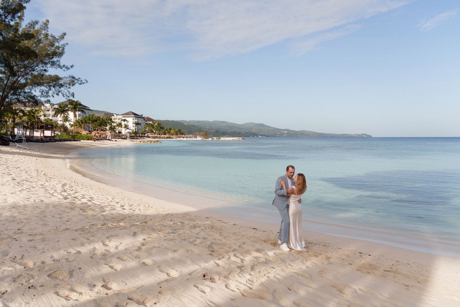 Couple in elegant attire standing together on tropical beach at sunset.