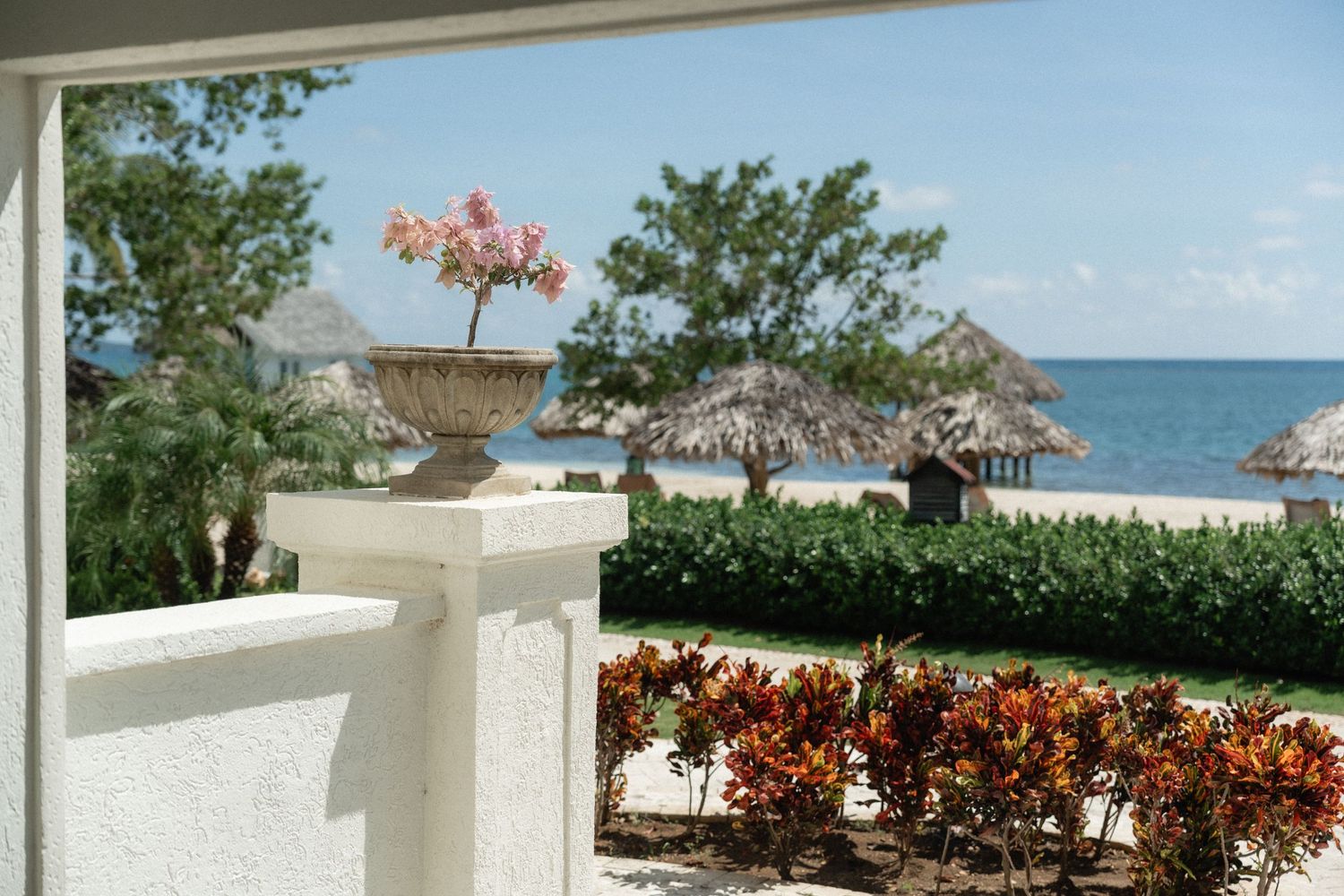 Ocean view from a white balcony with thatched beach huts and tropical flowers in the foreground.