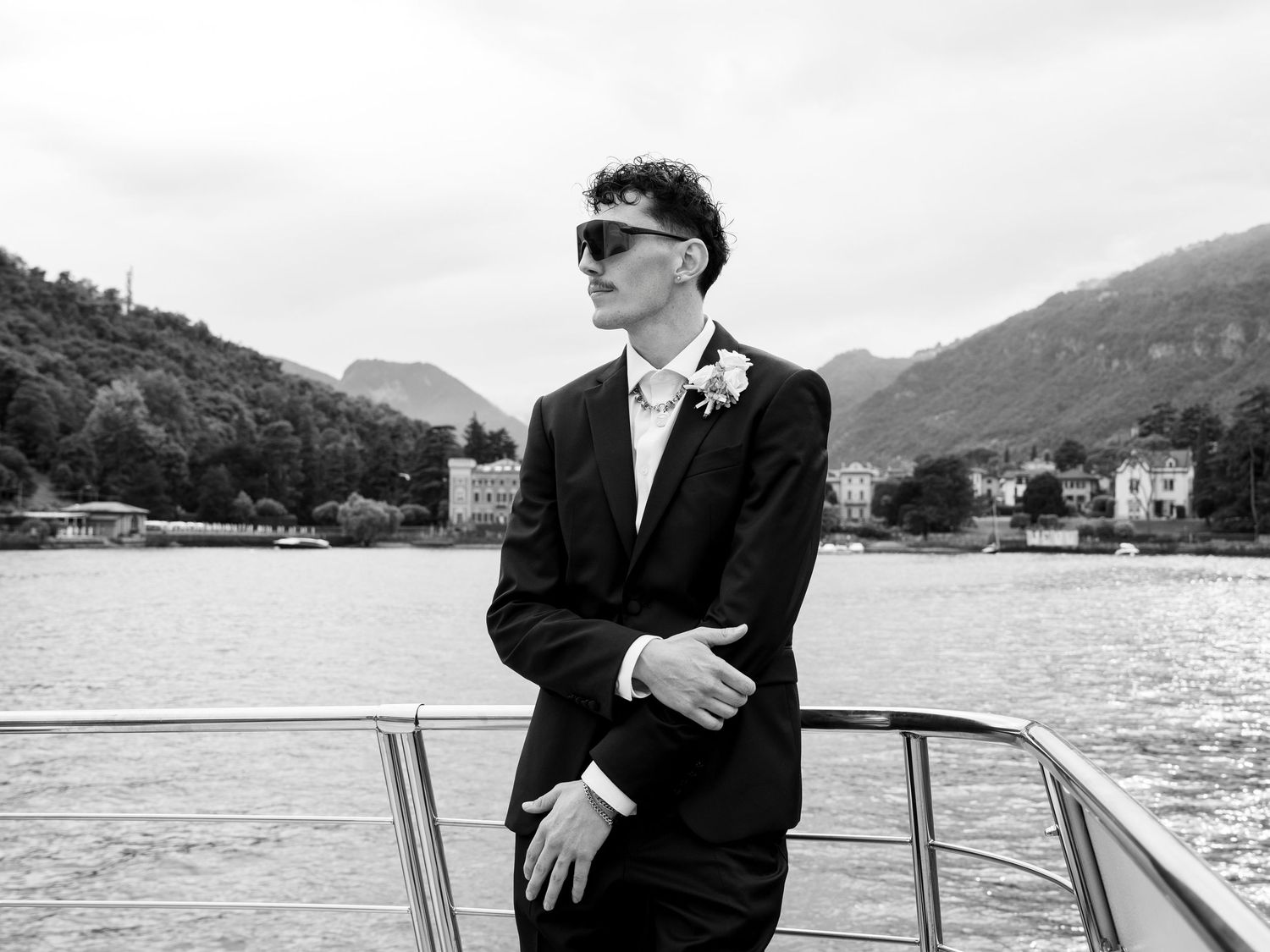 A person in a black suit stands on a boat with mountains and water in the background creating a scenic view.
