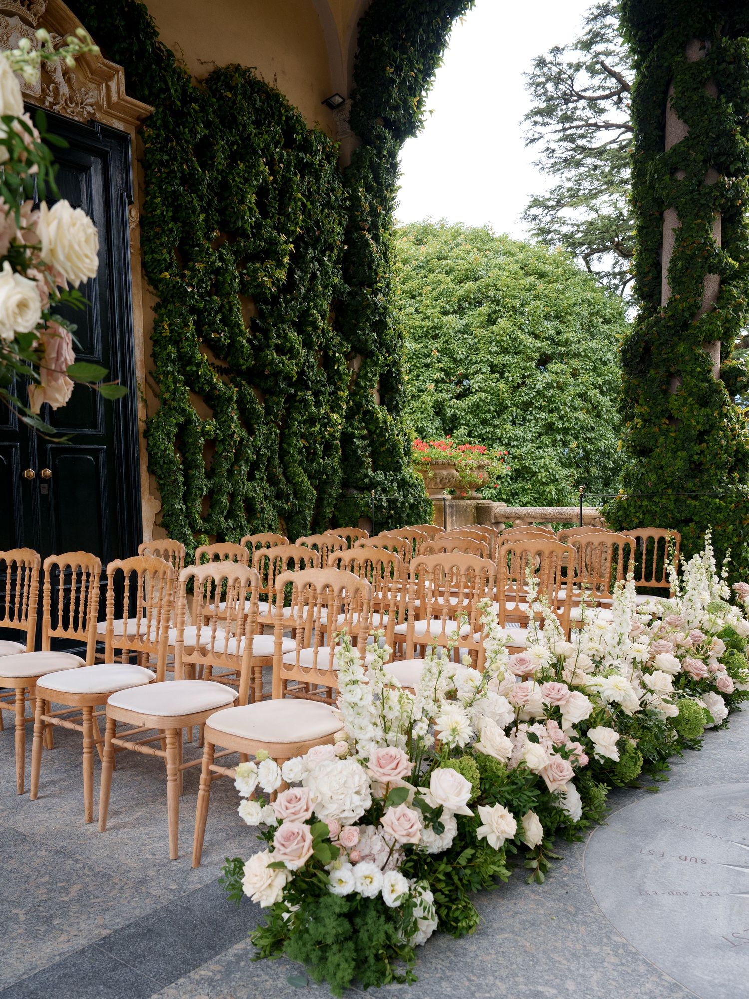 Elegant outdoor wedding ceremony setup with wooden chairs and lush white floral arrangements along a stone pathway.